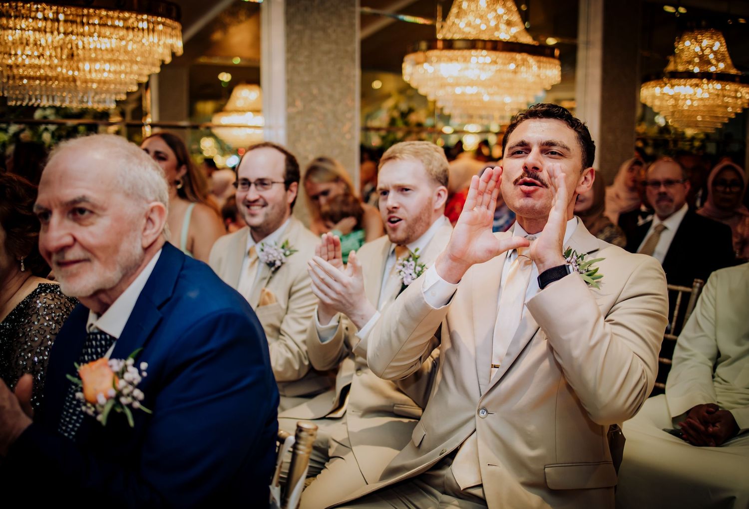 Wedding guests in light colored suits show emotional reactions during a ceremony under crystal chandeliers.