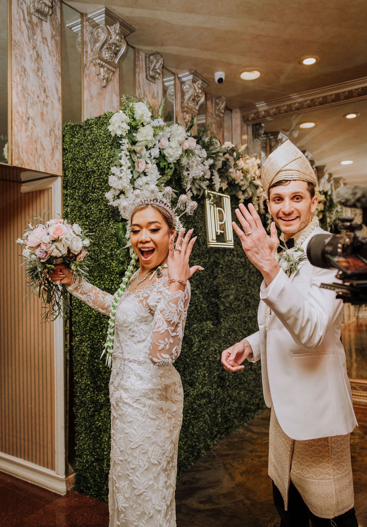 Newlyweds celebrate joyfully while showing off their rings in an elegantly decorated venue with greenery and floral decor.