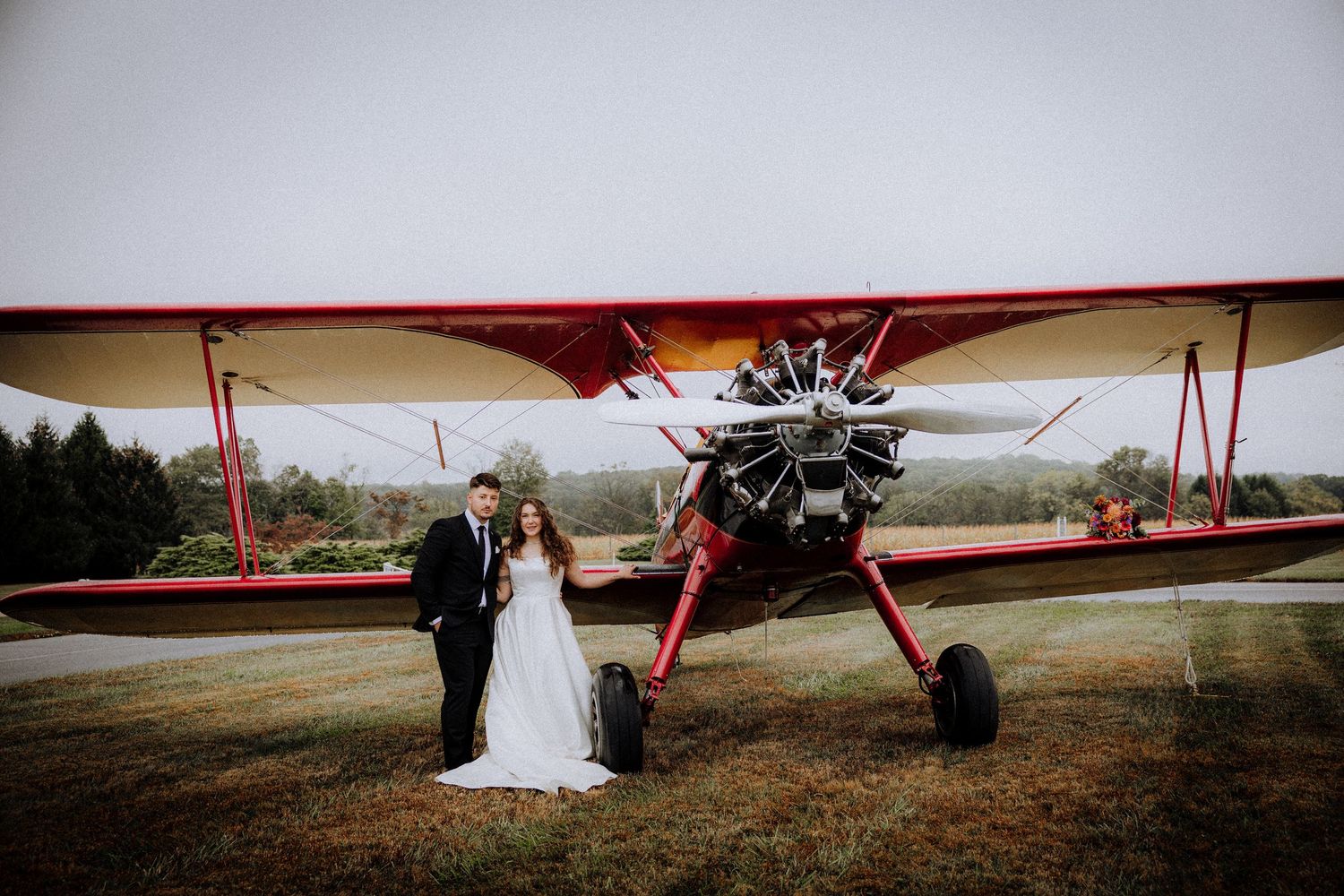 Wedding portraits with red vintage airplane in rustic outdoor setting.