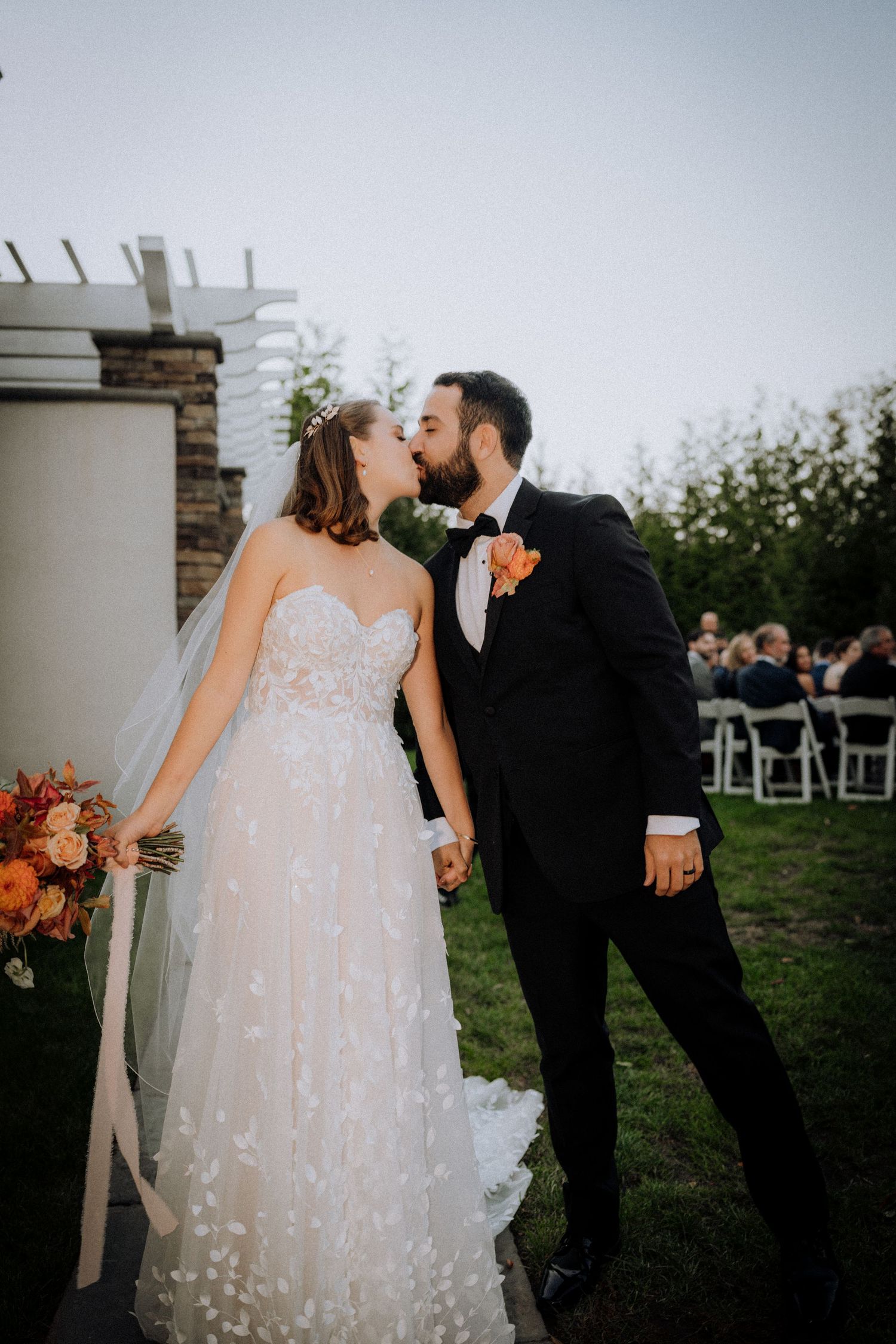 Newlyweds share an intimate moment outdoors near a white pergola with autumn flowers.