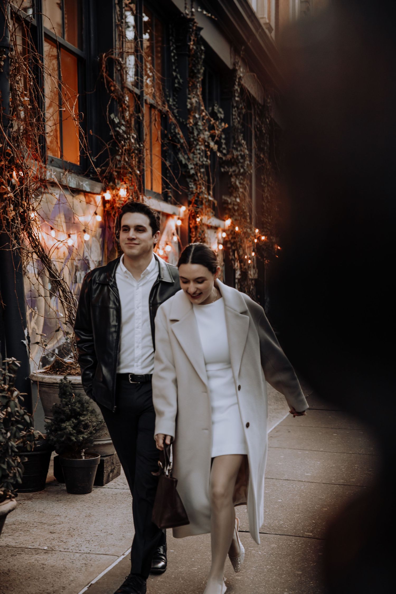 A stylish couple walks along a city sidewalk at dusk with twinkling lights and vines decorating the building facade.