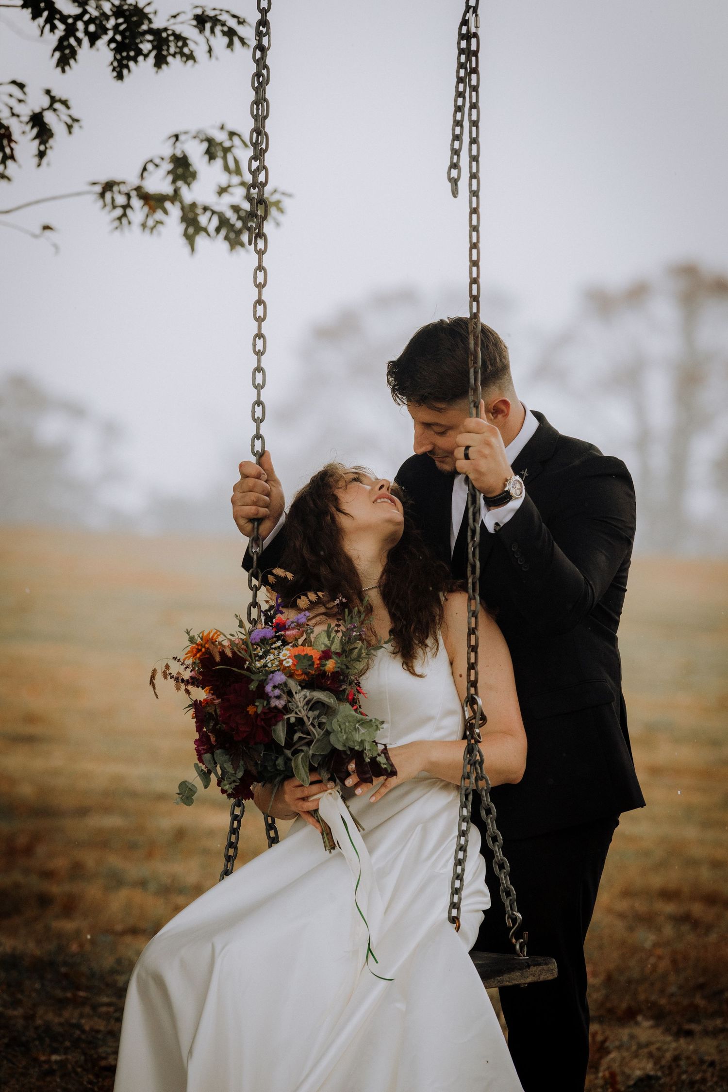 Newlyweds share intimate moments on a rustic swing set against a moody autumn landscape.
