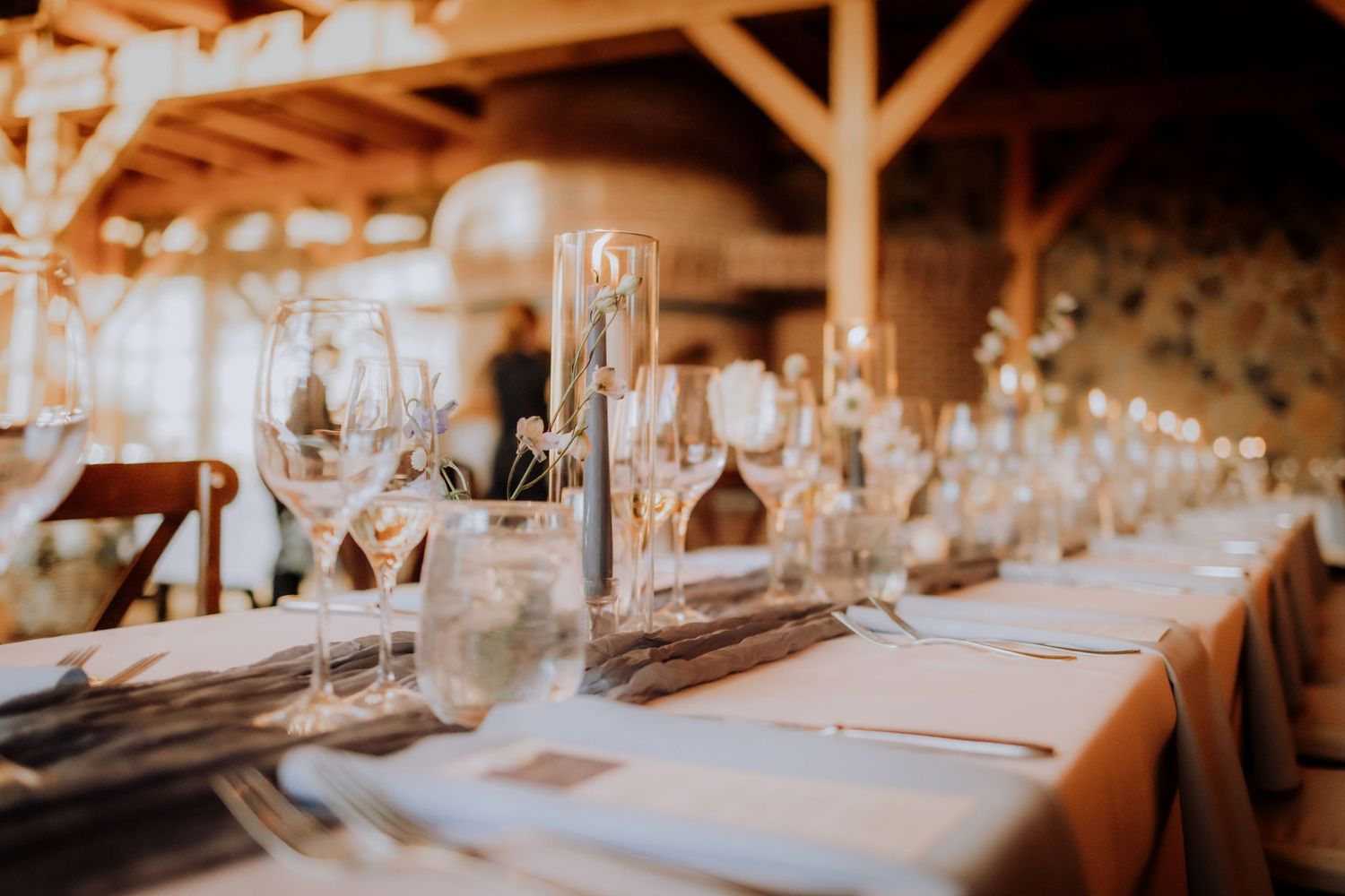Elegant rustic wedding reception table settings with glowing string lights and wine glasses in a barn venue.