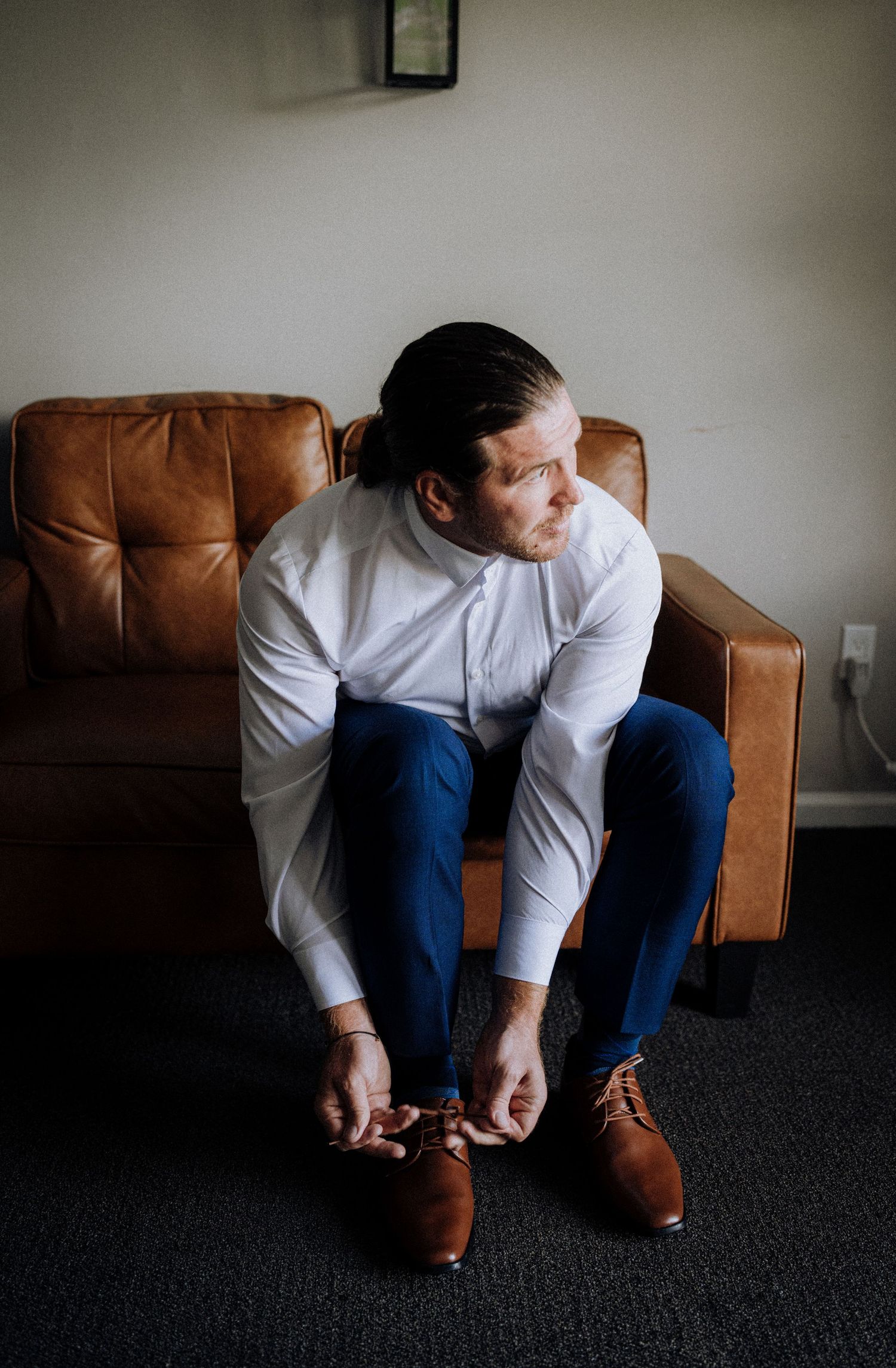 A person in formal attire sits on a brown leather couch while tying dress shoes.