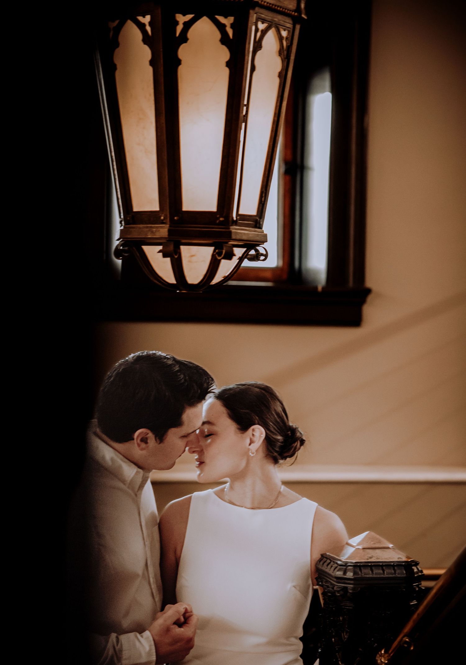Two people in elegant wedding attire share a tender moment in a warm-lit interior setting.