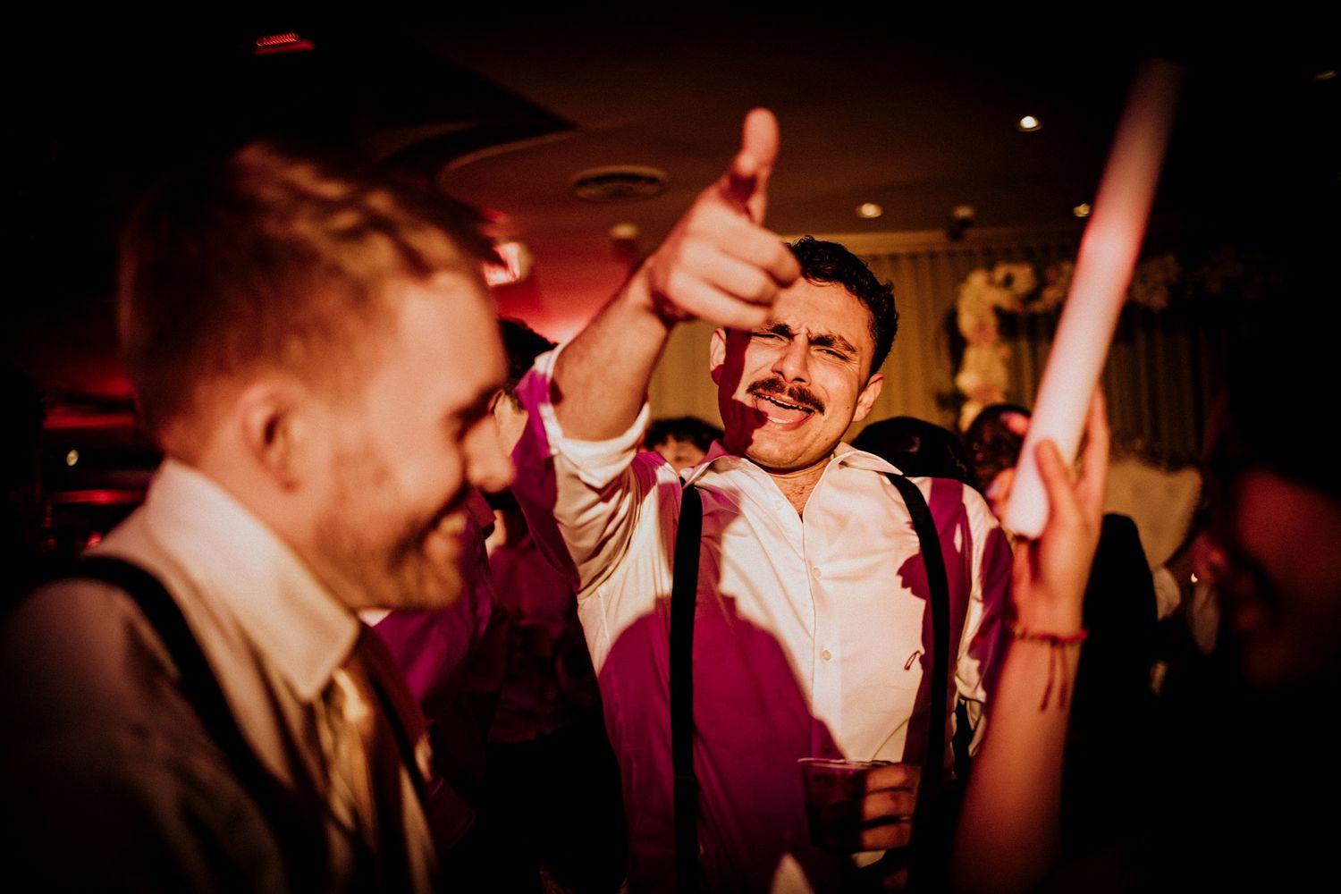 People dancing and celebrating at a lively wedding reception with red lighting and suspenders in focus.