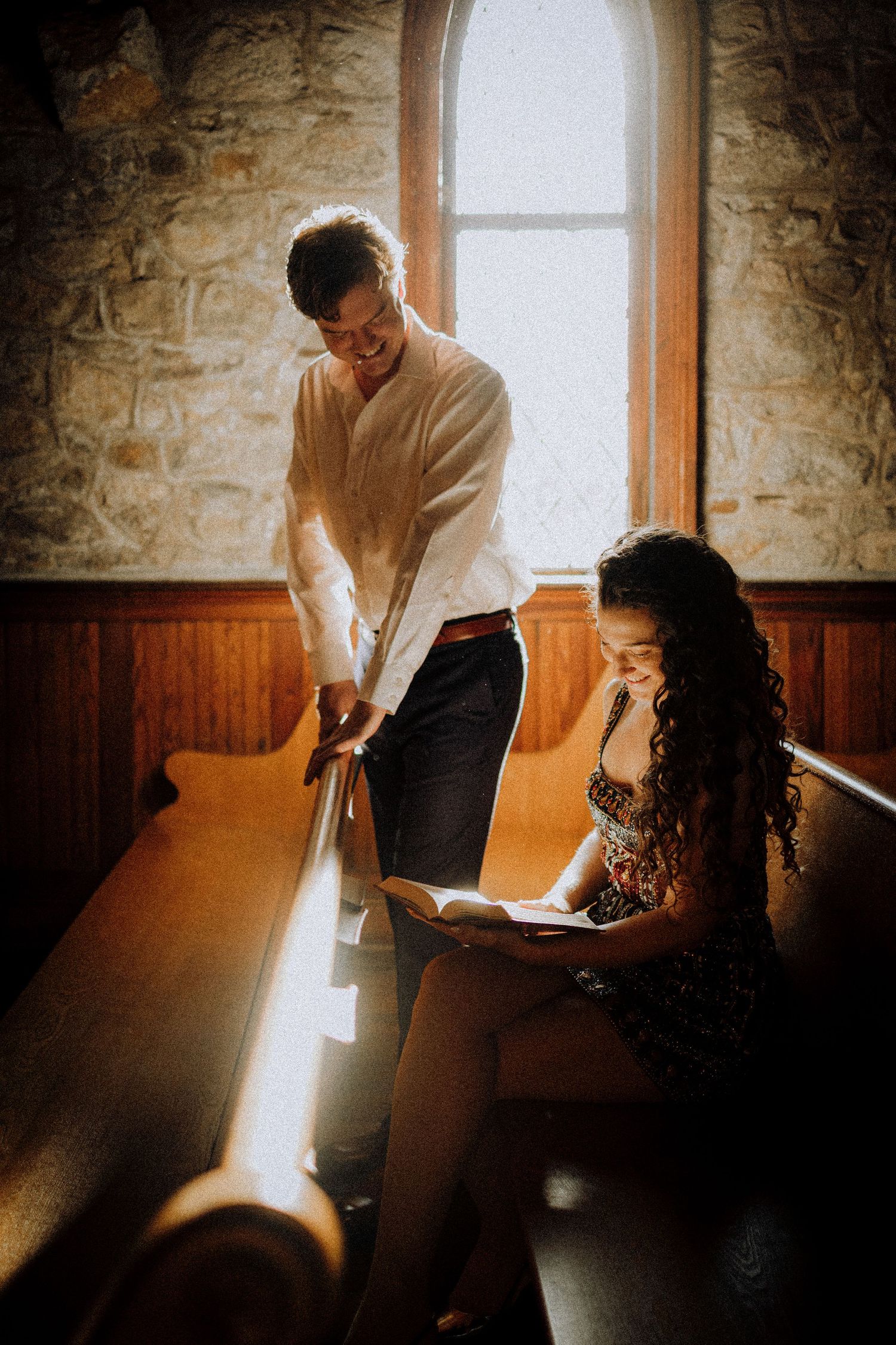 A couple in vintage attire sits in an old church pew bathed in natural window light.