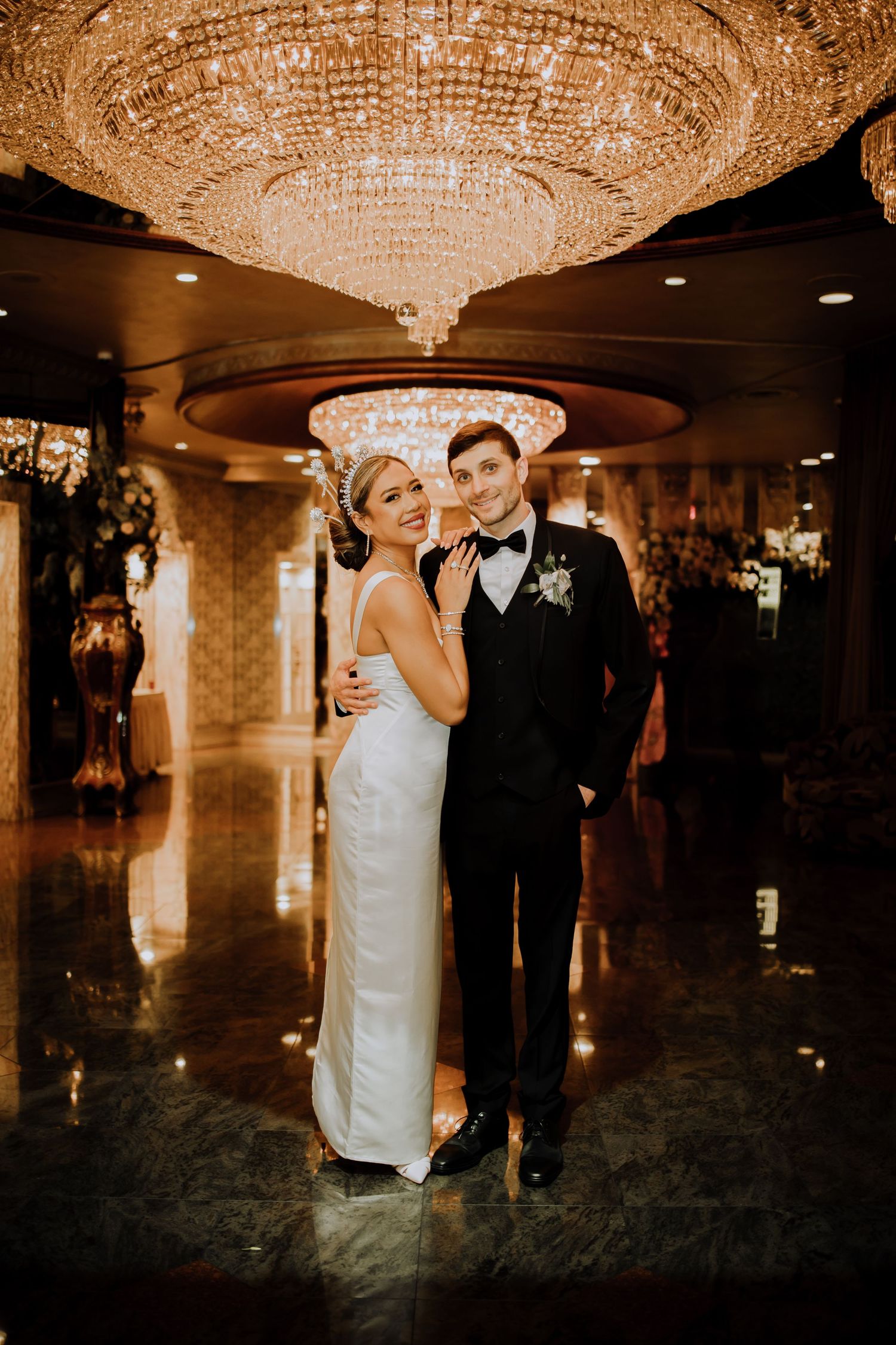 A couple dancing romantically beneath a grand crystal chandelier at an elegant ballroom reception venue.