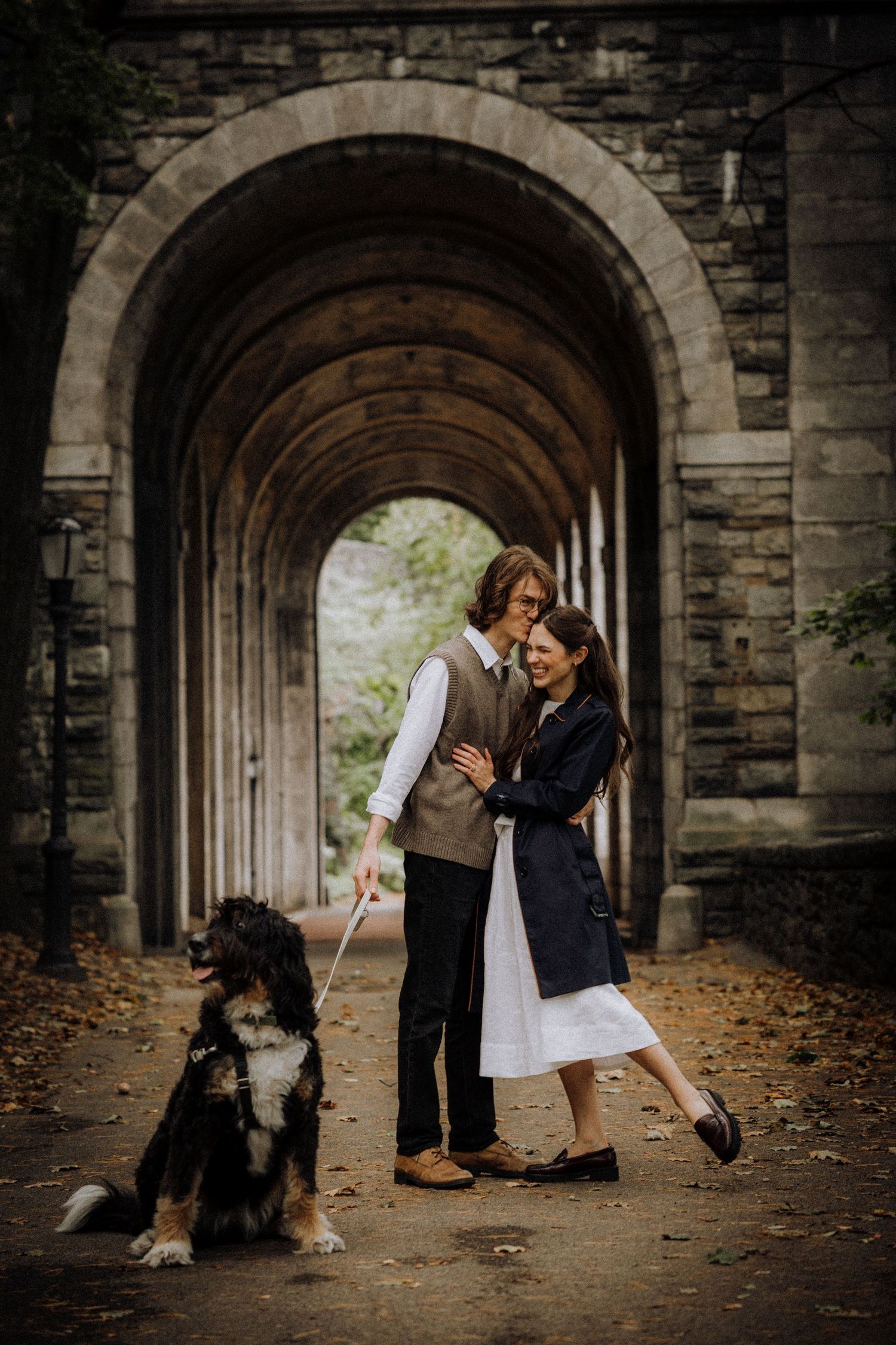 A couple and their dog pose together under a beautiful stone archway on an autumn day.