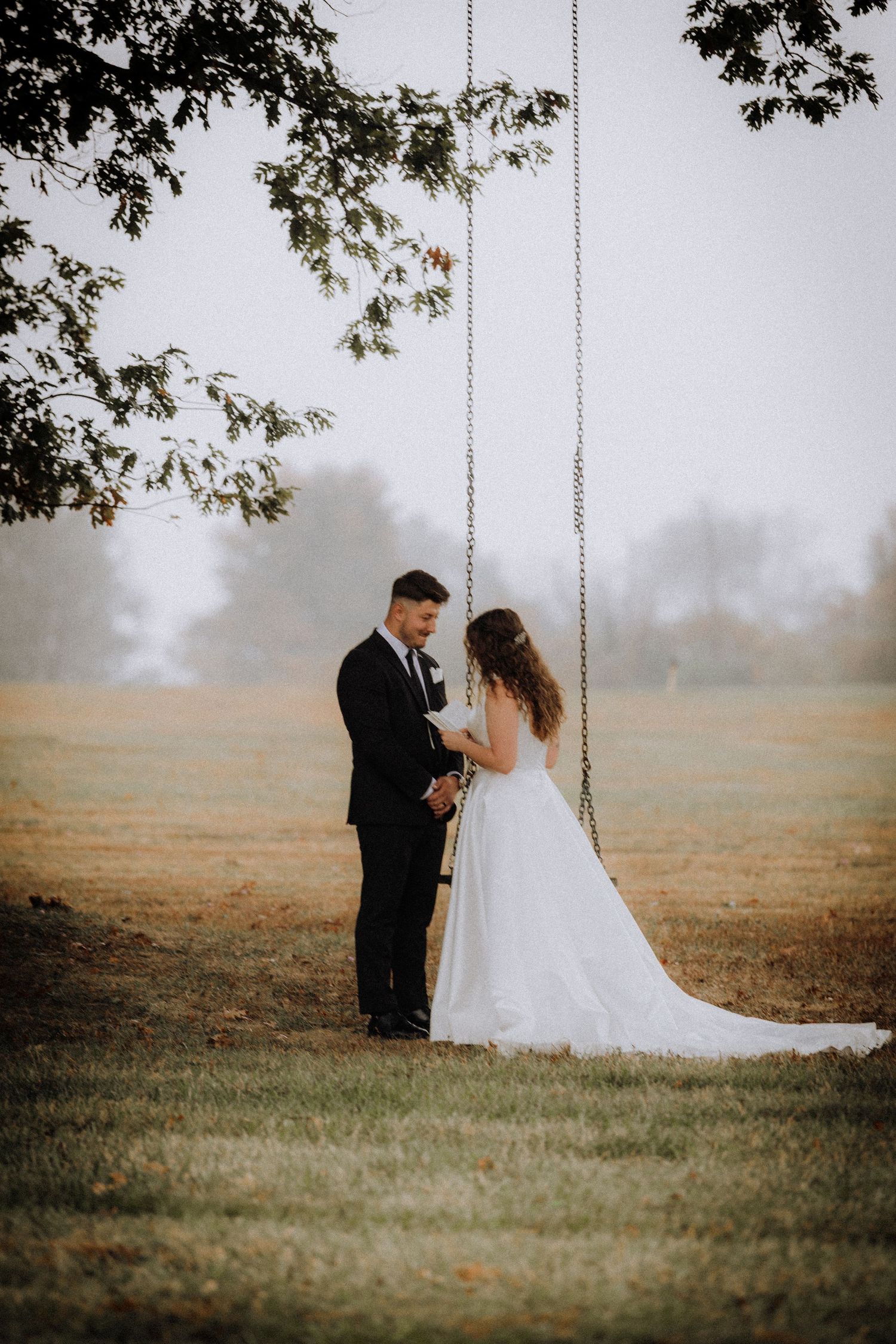 A romantic scene with a couple near tree swings in a misty field during golden hour.