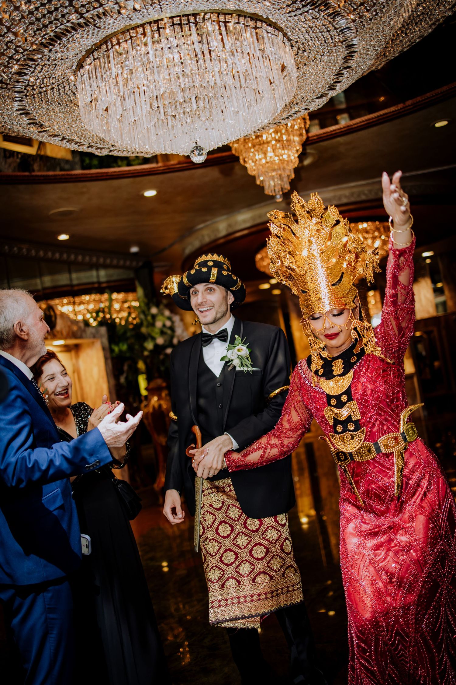Dancing figures celebrate at an elegant reception under crystal chandeliers during what appears to be a formal event.