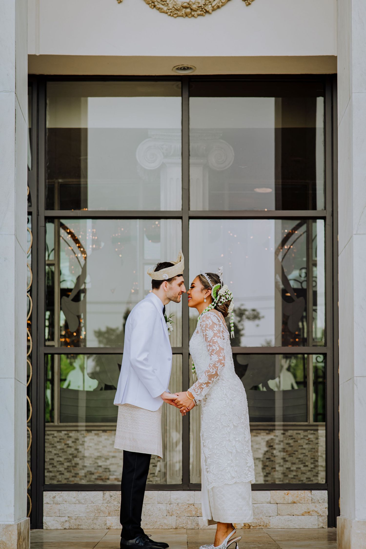A couple in white attire share a tender moment in front of glass doors at an elegant venue entrance.
