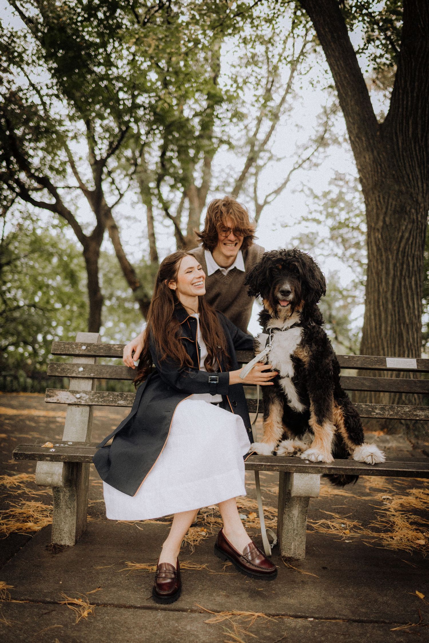 A couple sits on a park bench with their large fluffy dog on an autumn day.