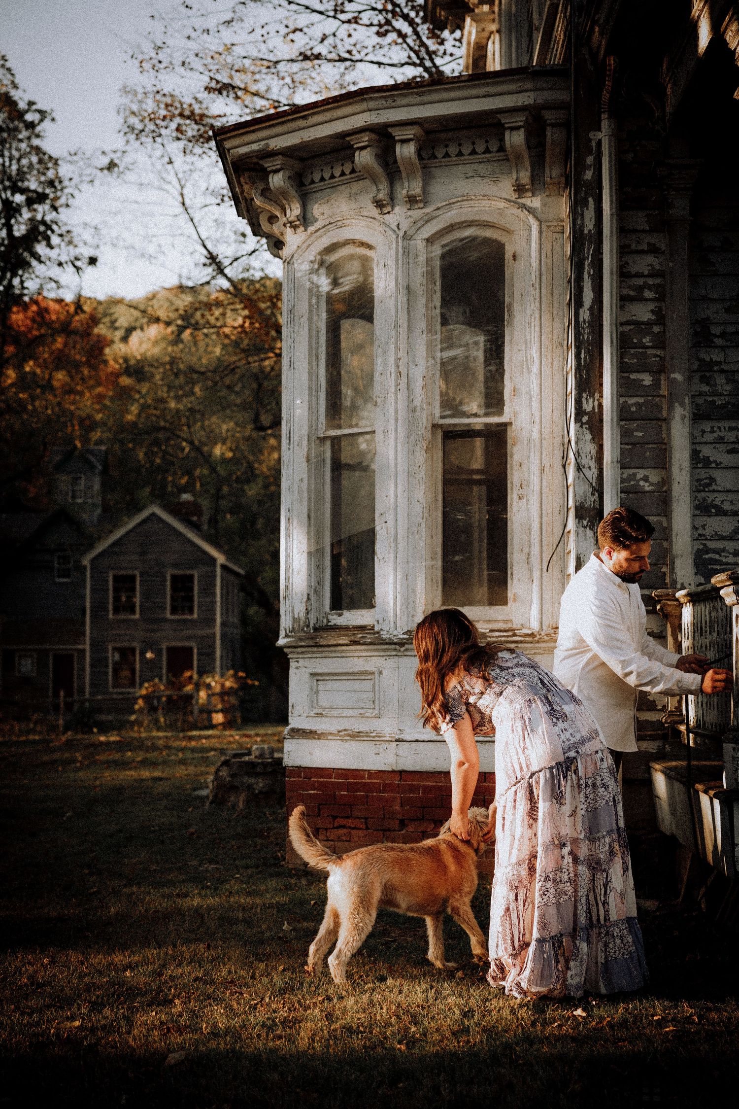 A couple and their dog explore the porch of a weathered Victorian home at sunset.