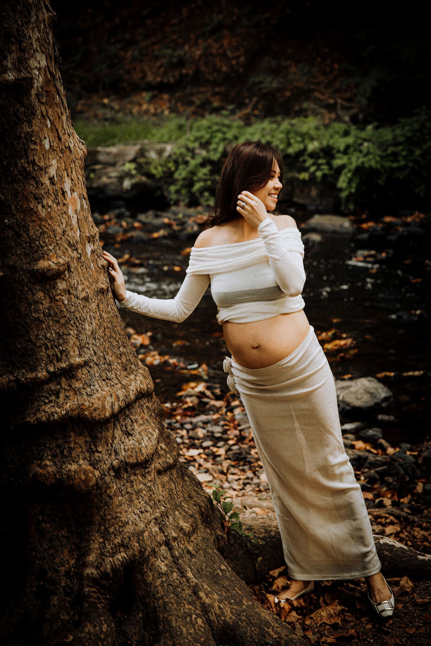 Artistic maternity portraits against a large tree trunk with scattered autumn leaves on the ground.