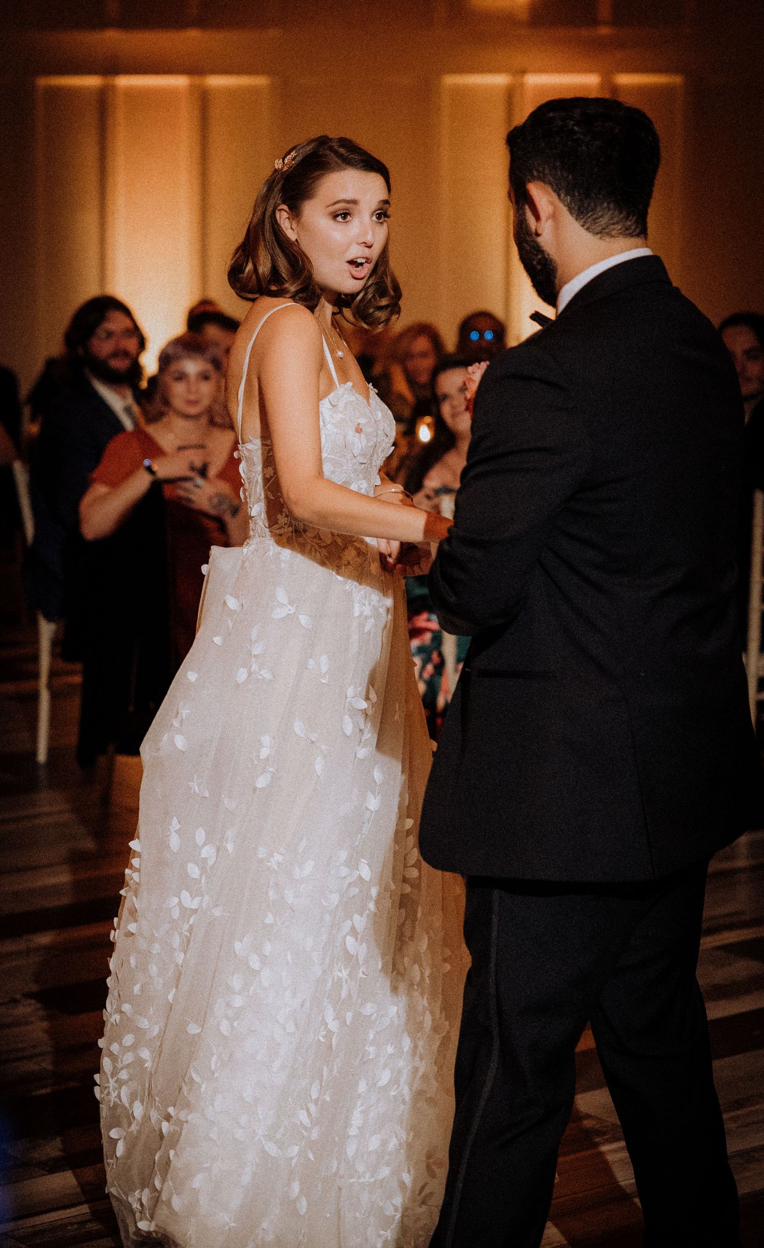 Couple dances in elegant ballroom with bride wearing flowing white butterfly detailed gown.