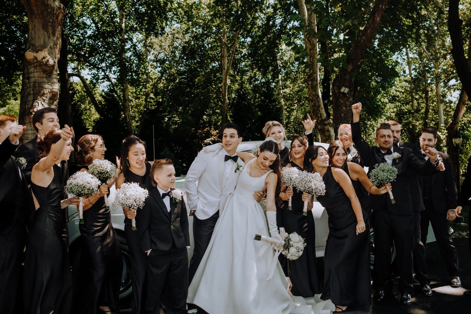 Wedding guests in black attire gather outdoors under trees for a celebration.