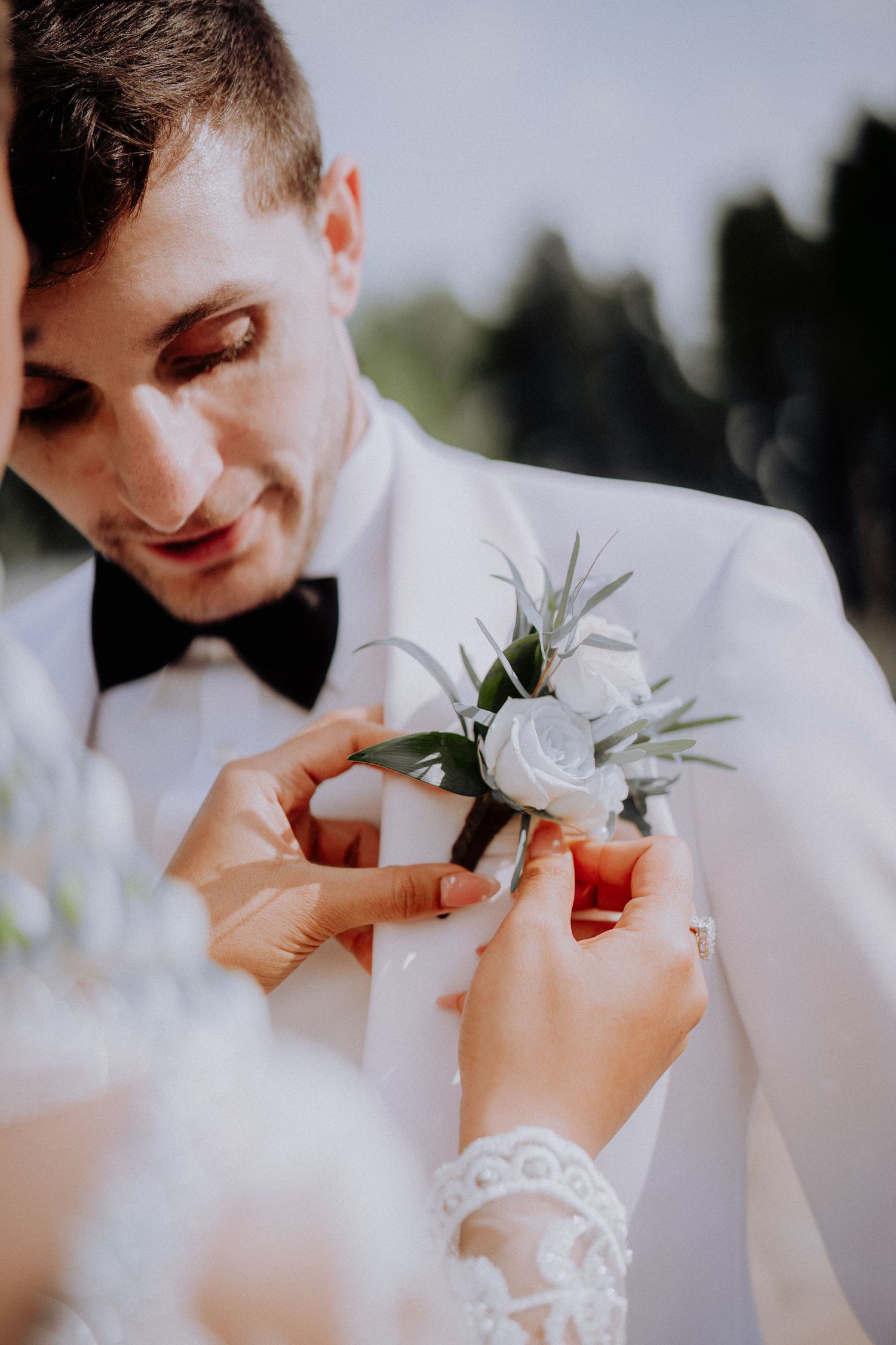 Hands adjusting a delicate white rose and greenery boutonniere on a crisp white tuxedo jacket at a wedding ceremony.