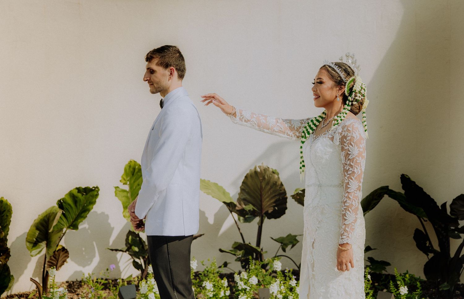 A couple in wedding attire poses against a white wall with tropical plants, the bride reaching toward the groom.