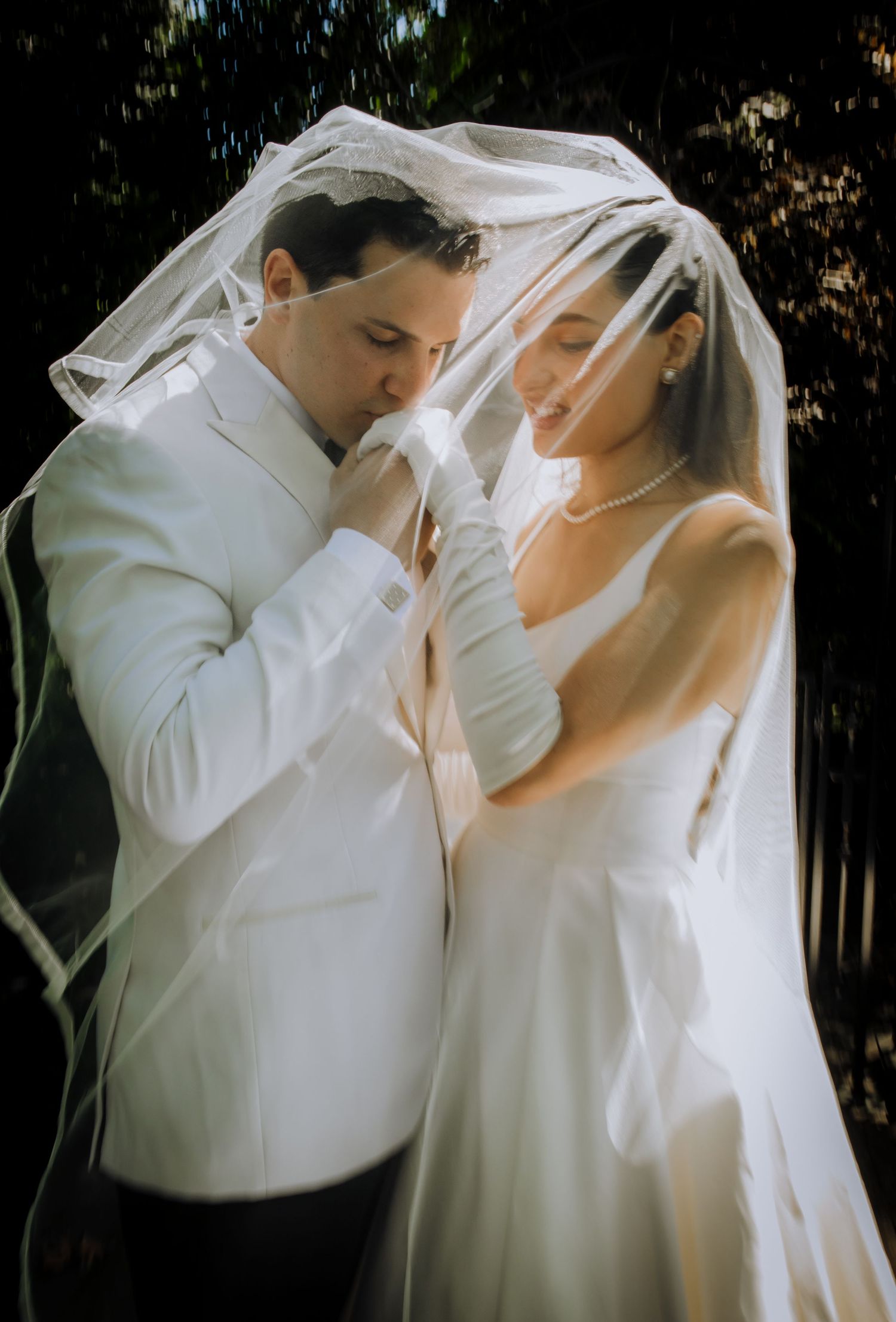A romantic wedding moment captured under a flowing white veil as a couple shares an intimate gesture outdoors.