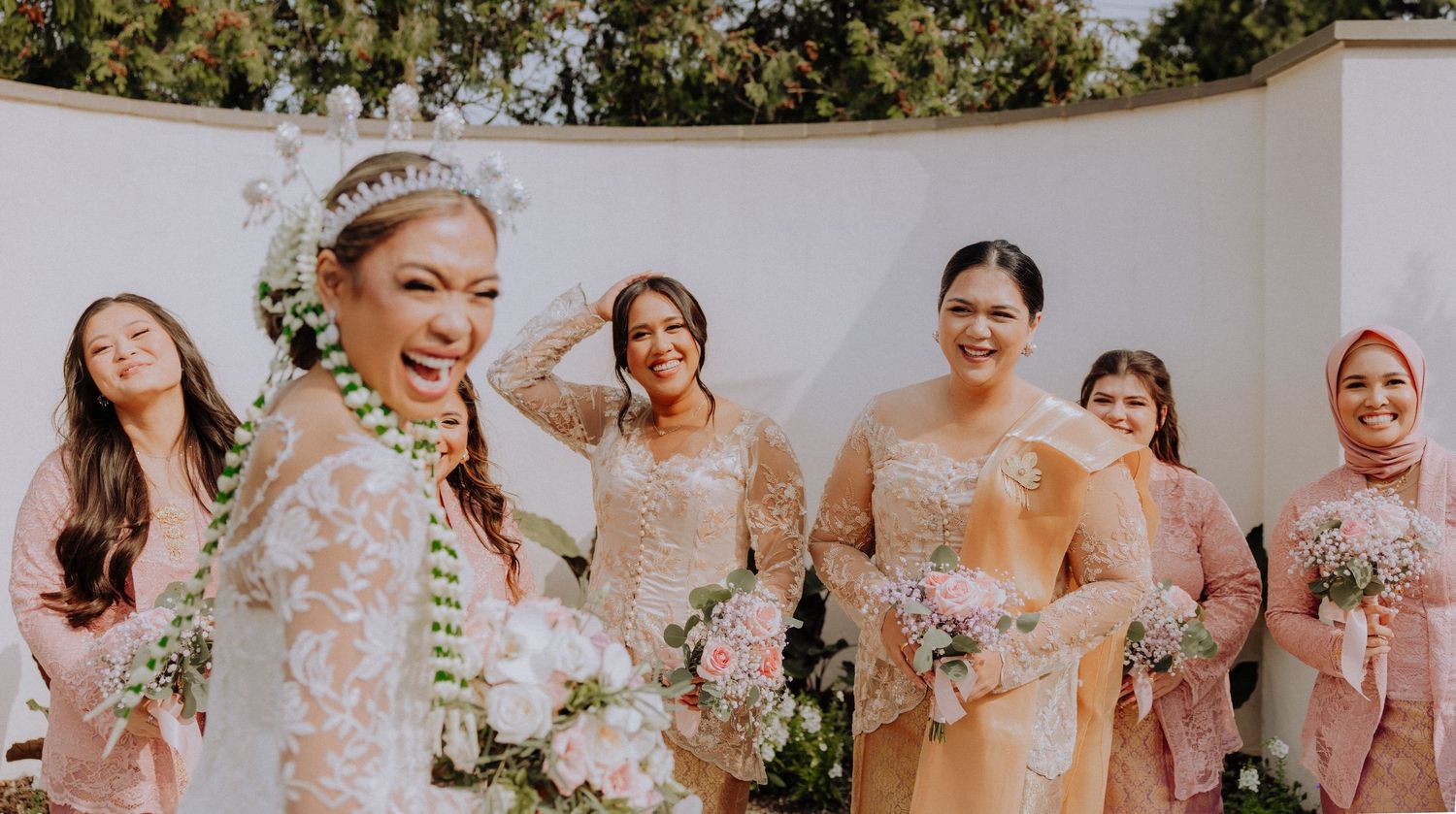 A bridal party in soft pink and neutral dresses laughs together while holding white floral bouquets during wedding photos.