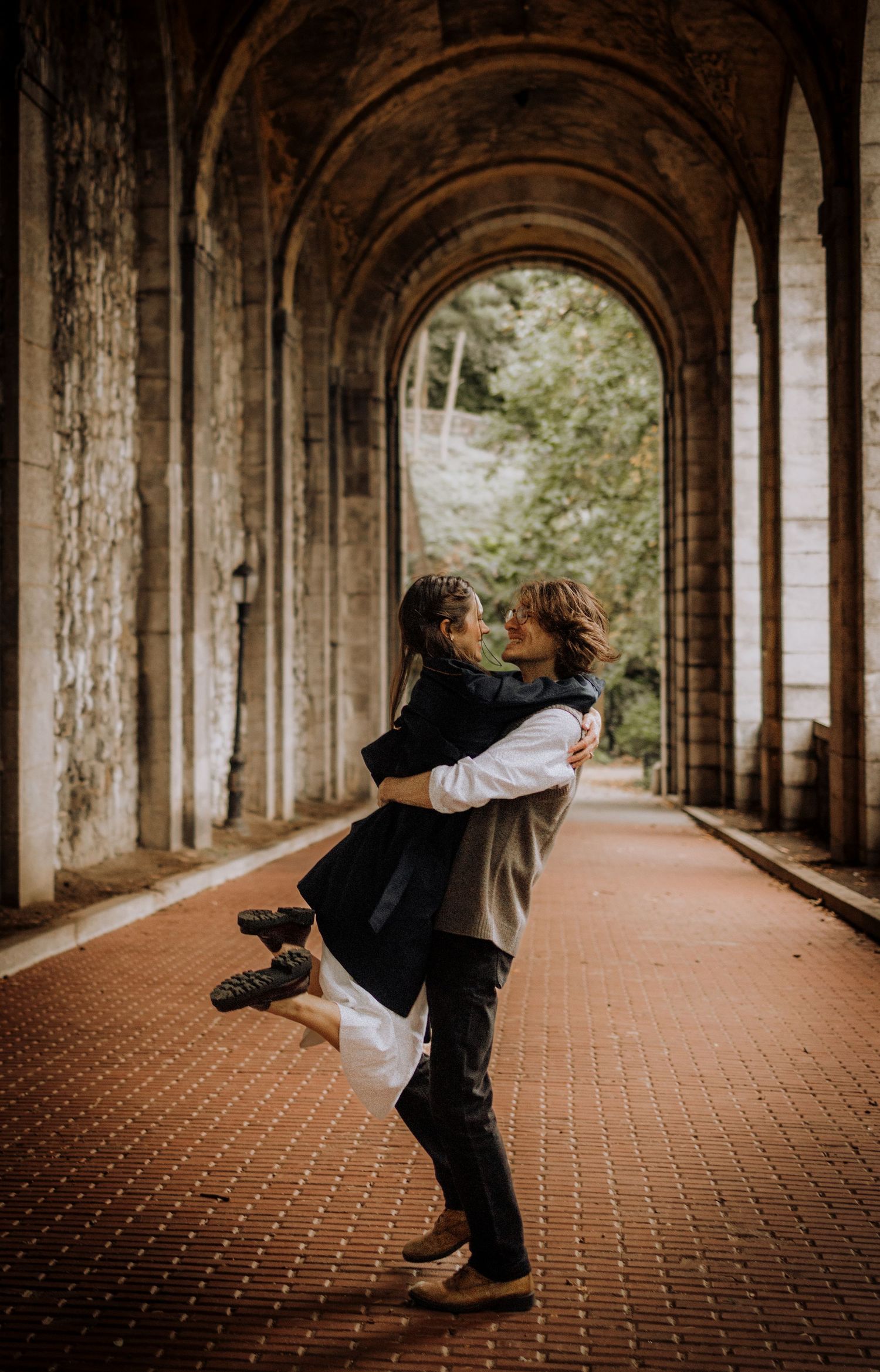 A couple dances playfully in an arched brick corridor with dramatic lighting and architectural columns.