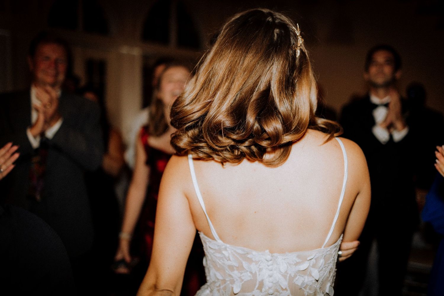 Back view of a bride in a white lace dress with guests clapping during a wedding reception.
