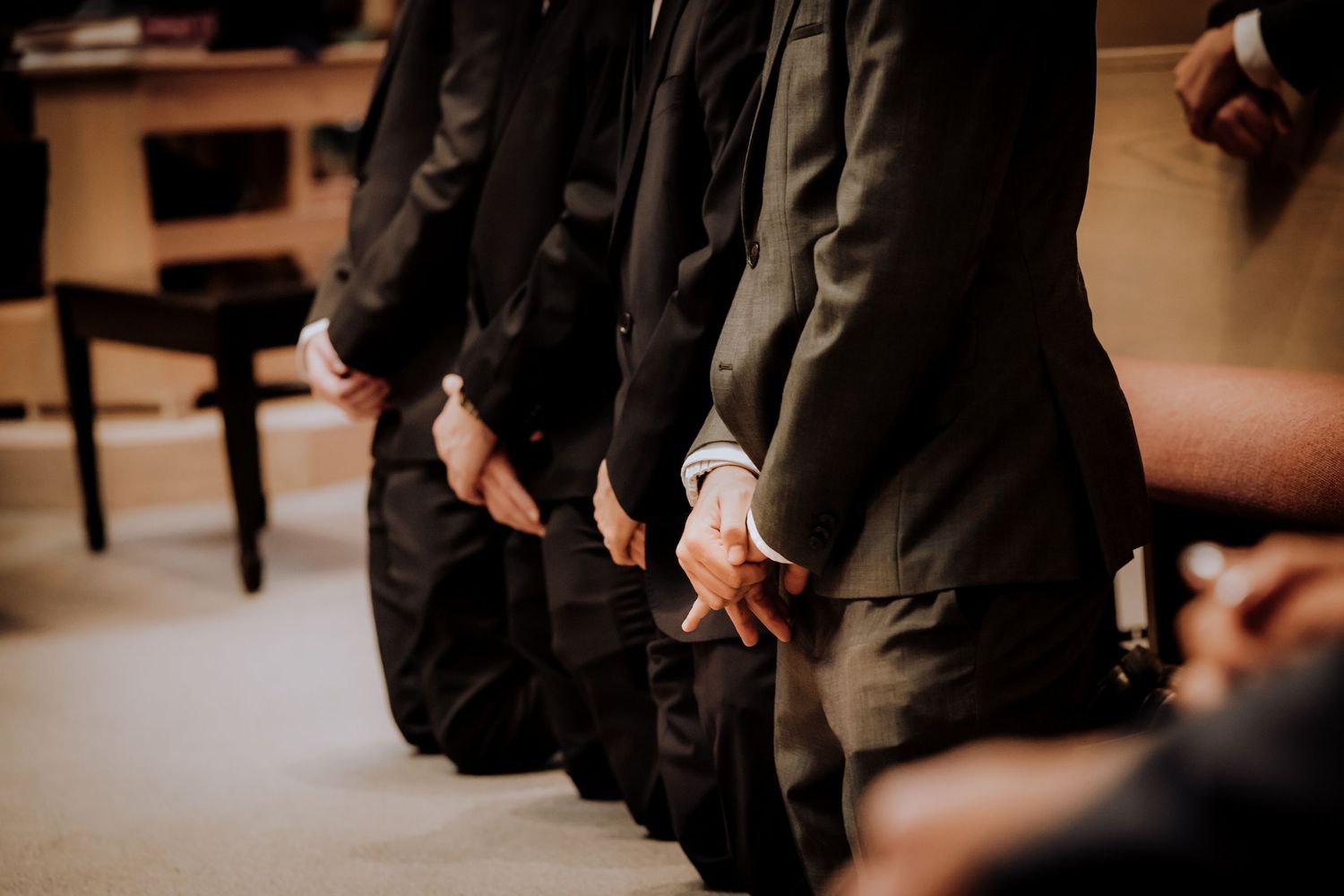 People in dark formal attire kneeling in prayer or meditation during a ceremony in a dimly lit room.