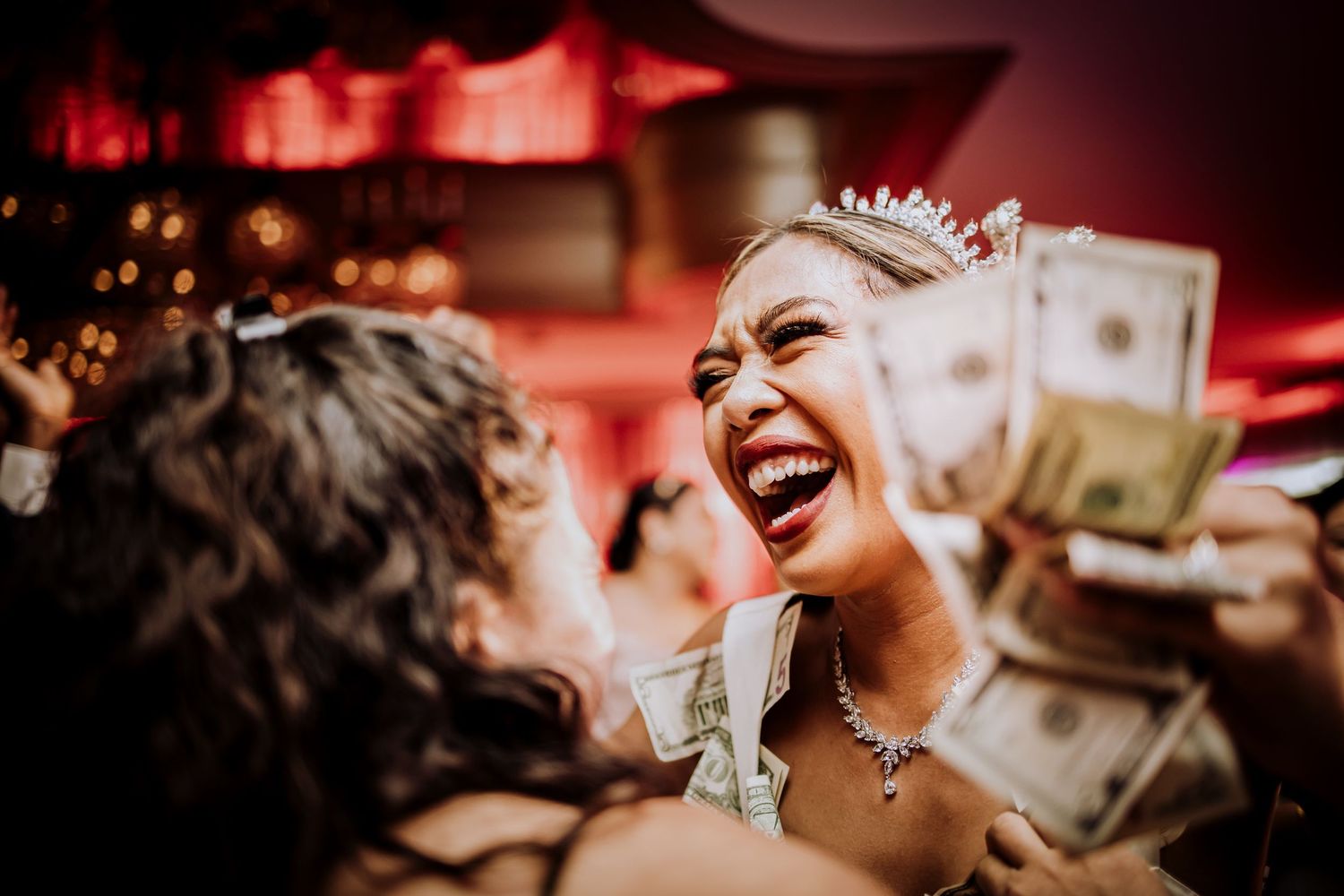 A candid wedding reception moment showing laughter and money being thrown in the air at a party with red ambient lighting.