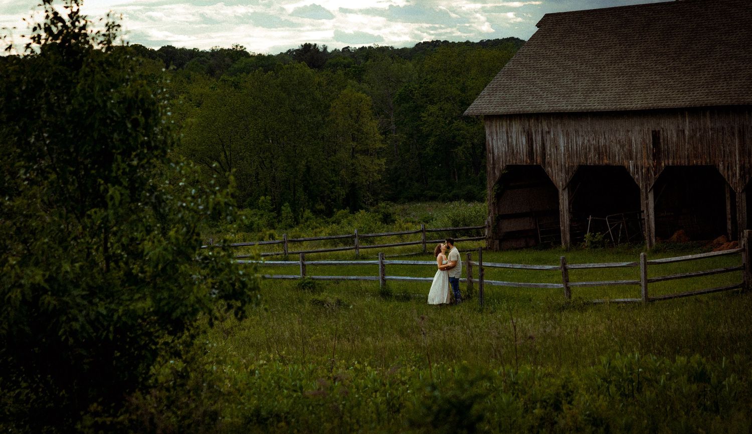 A couple in formal attire stands near a rustic wooden barn in a lush green meadow at sunset.