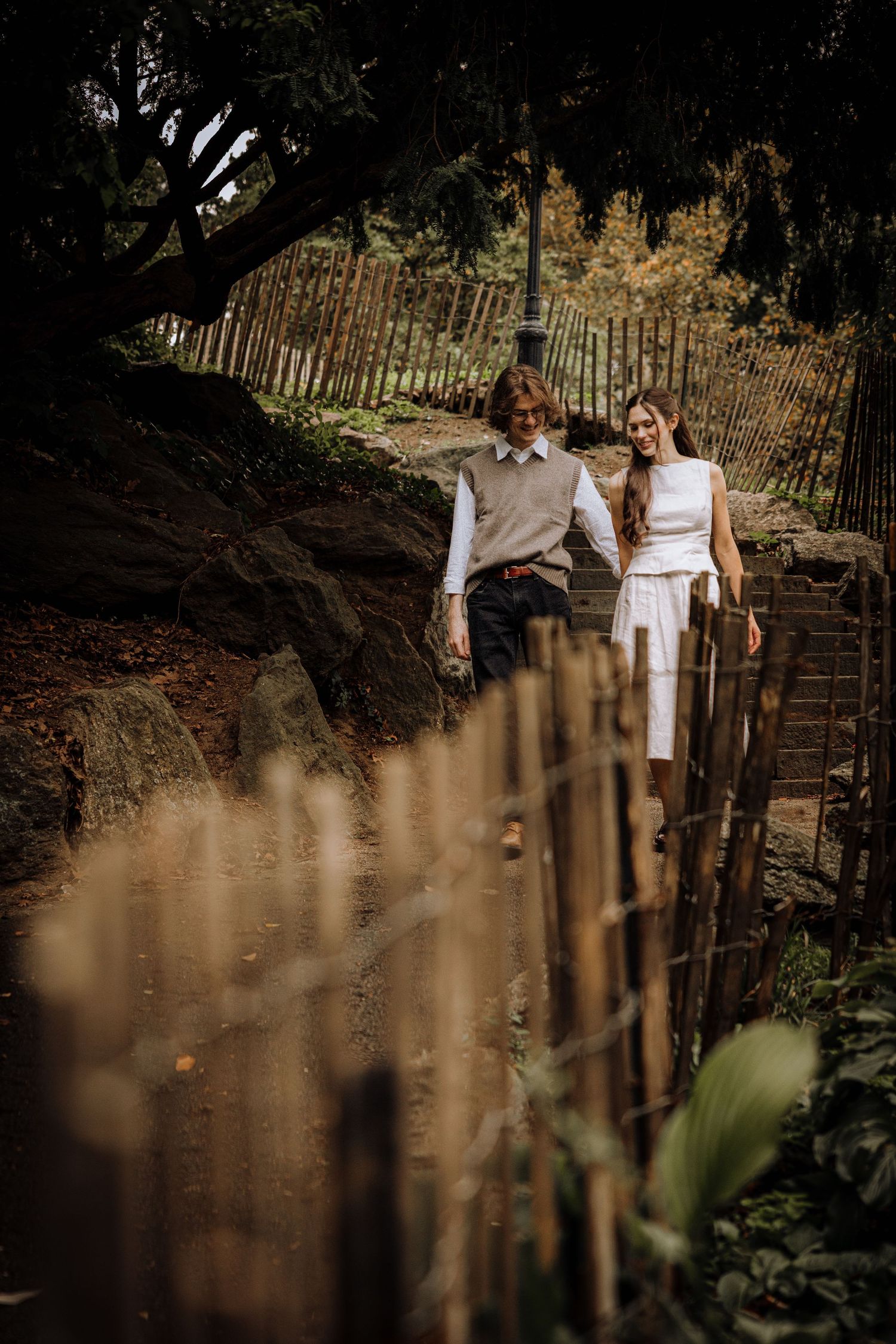 A couple stands together on rustic wooden steps along a weathered fence in a moody, artistic outdoor setting.