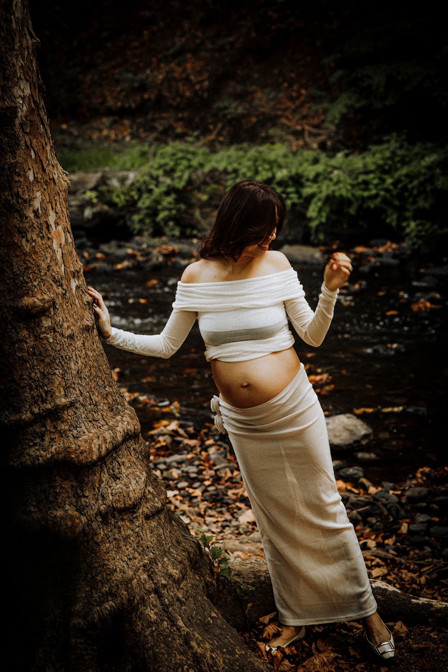 Artistic maternity photos showing a white dress against dark tree trunks and fallen autumn leaves.