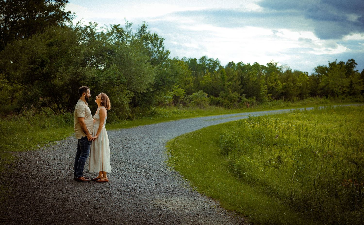 Nature path engagement session with dramatic cloudy sky and green meadow backdrop.