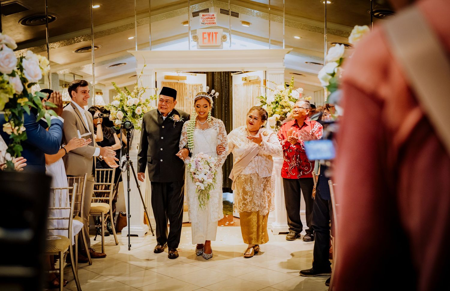 Wedding celebration with a group of people walking down an illuminated indoor venue hallway with floral decorations.