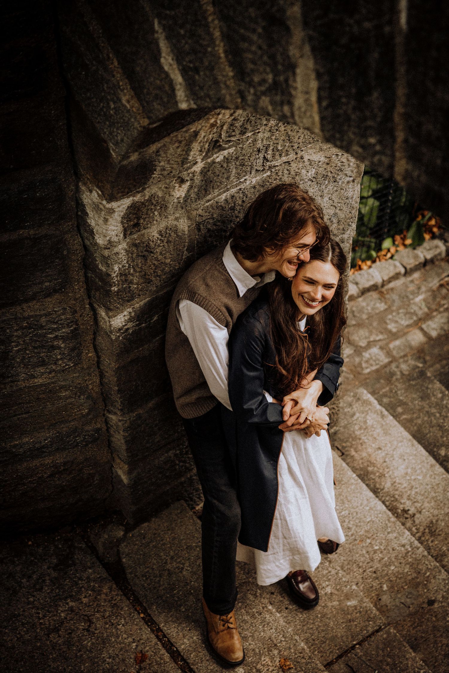 A romantic couple embraces and laughs together against a rustic stone wall backdrop in warm lighting.