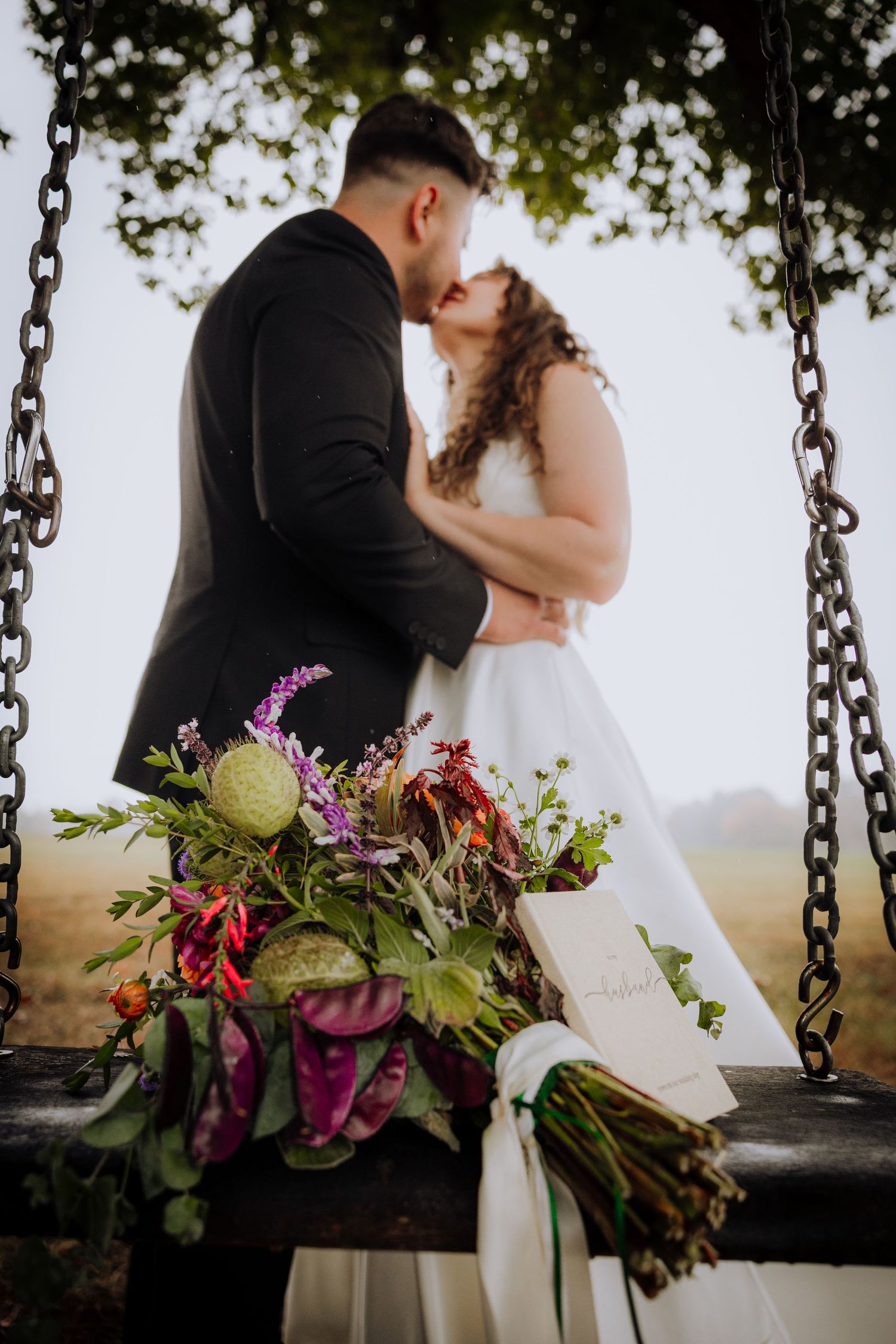 Romantic wedding couple sitting on a rustic swing adorned with colorful wildflower bouquet at sunset.