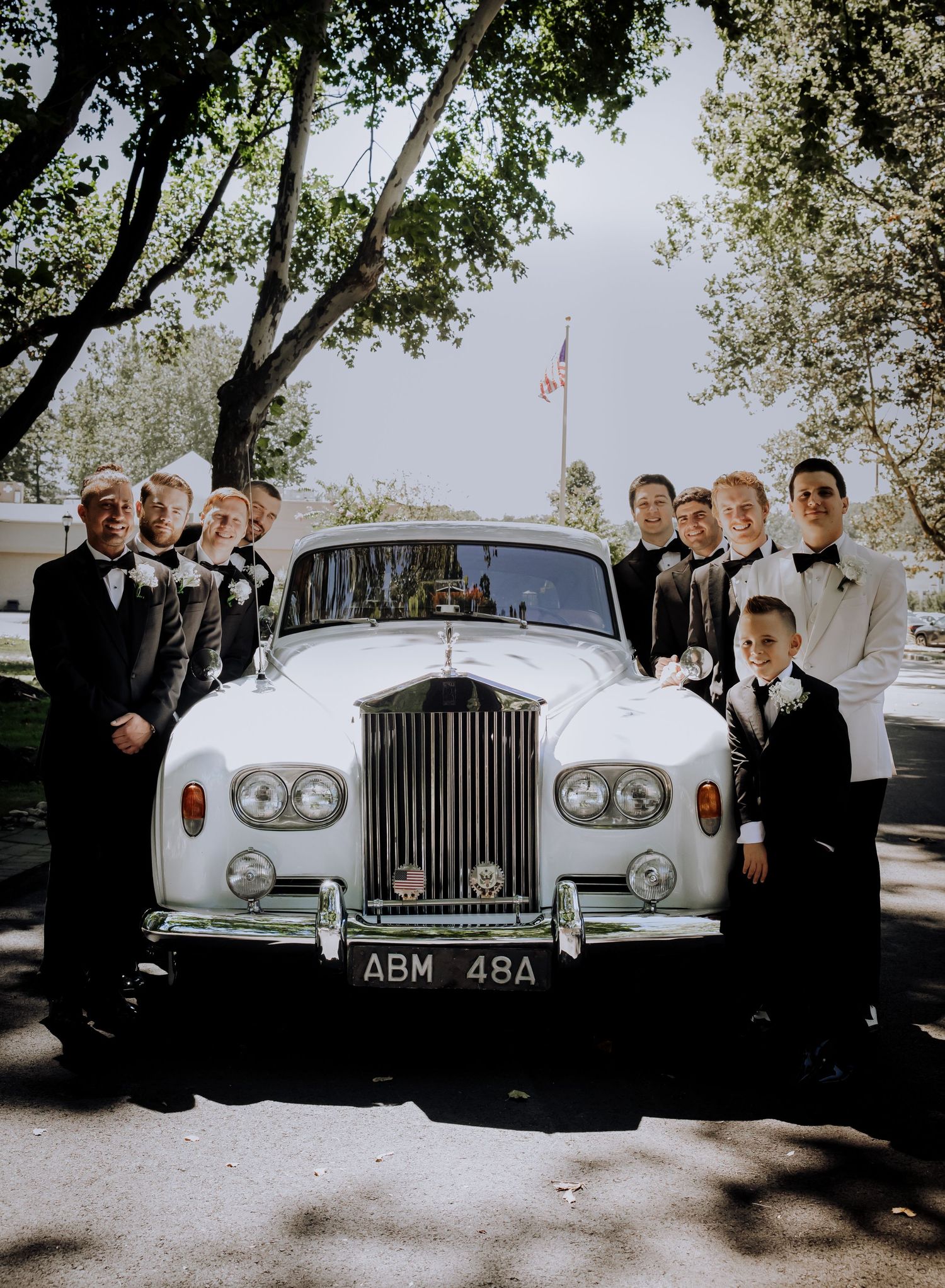 A classic silver Rolls Royce wedding car surrounded by wedding party members in formal attire.
