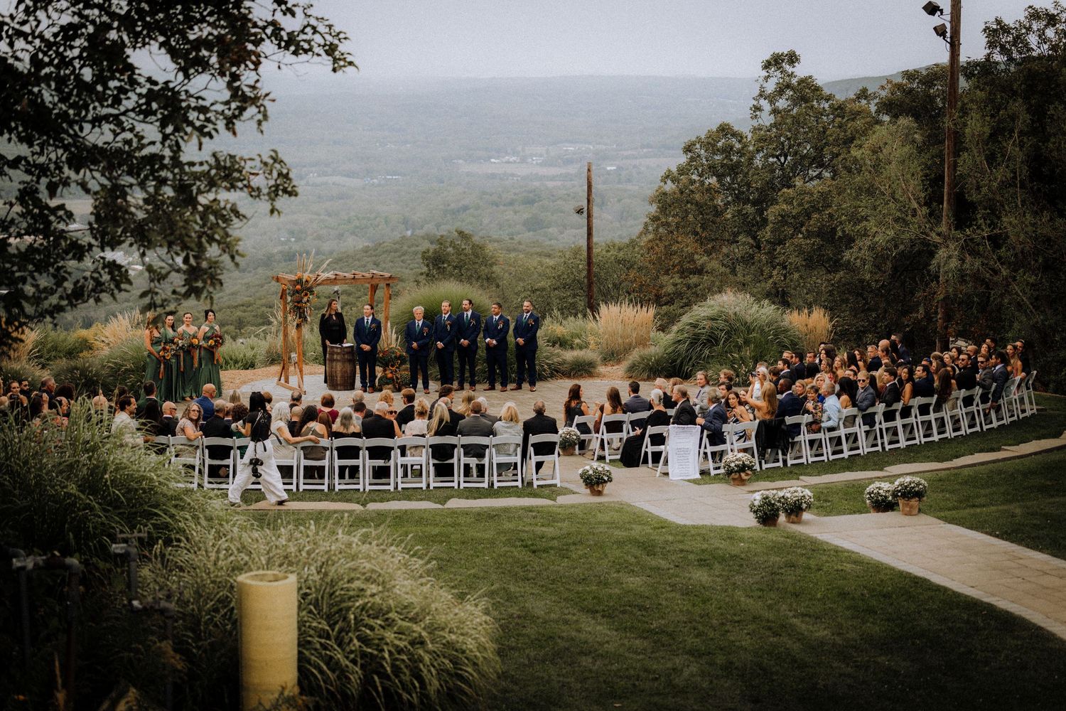 Scenic outdoor wedding ceremony setup with guests seated on a hilltop overlooking a valley.