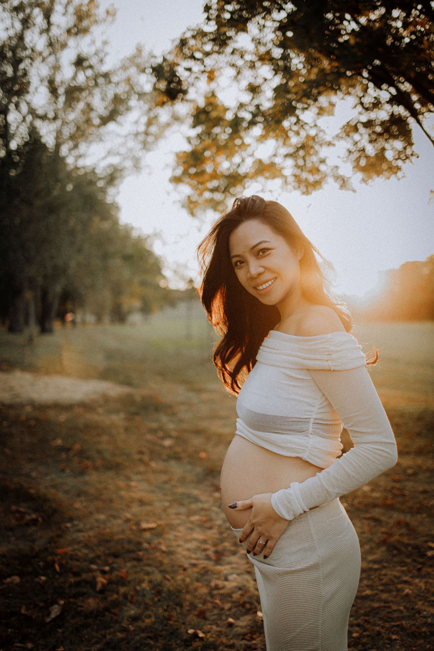 Expectant mother in white dress poses during golden hour outdoor photoshoot.
