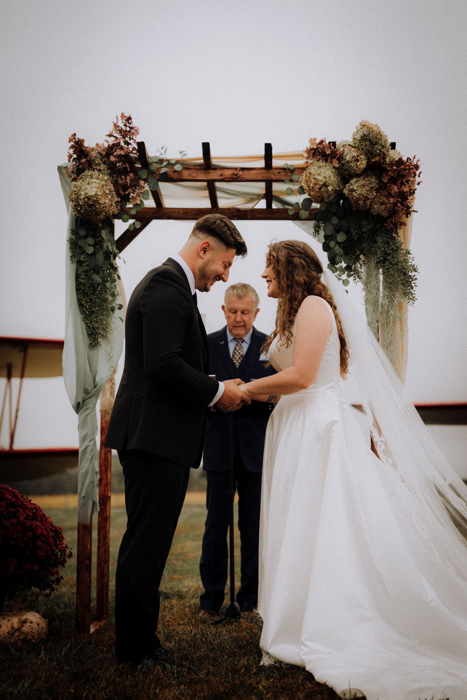 A romantic outdoor wedding ceremony takes place under a rustic wooden arch adorned with dried flowers and greenery.