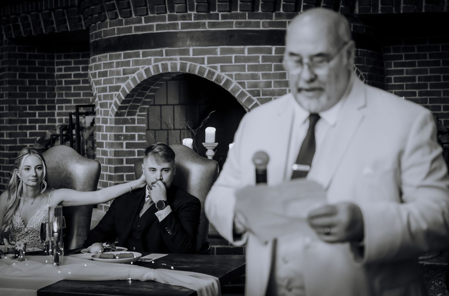 Black and white panoramic photo captures candid moments during wedding reception toasts at brick venue.