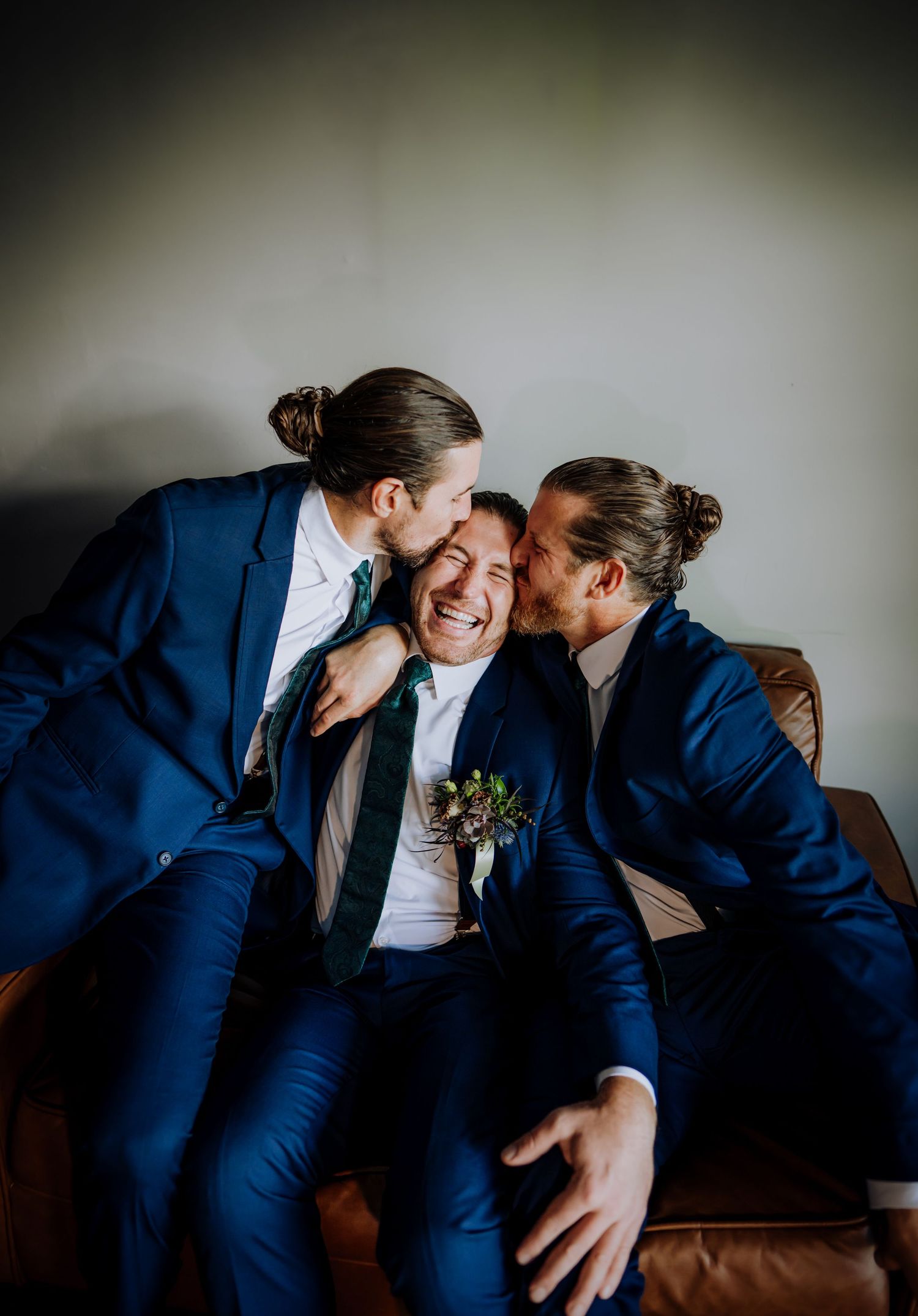 Wedding party members in navy suits share a candid laughing moment on a leather couch.