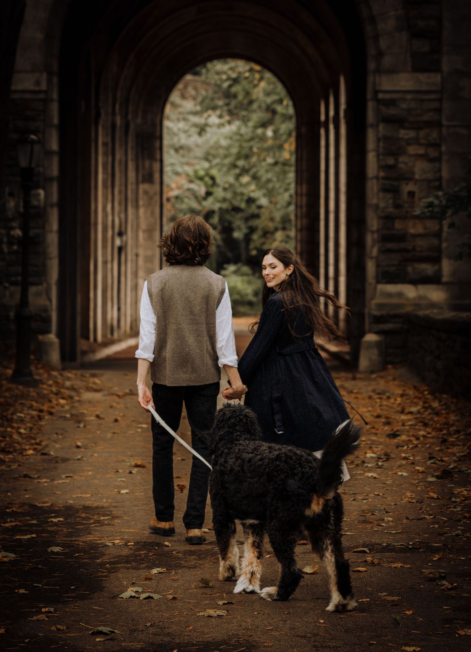A couple walks with their black dog through a stone archway covered in fallen leaves.