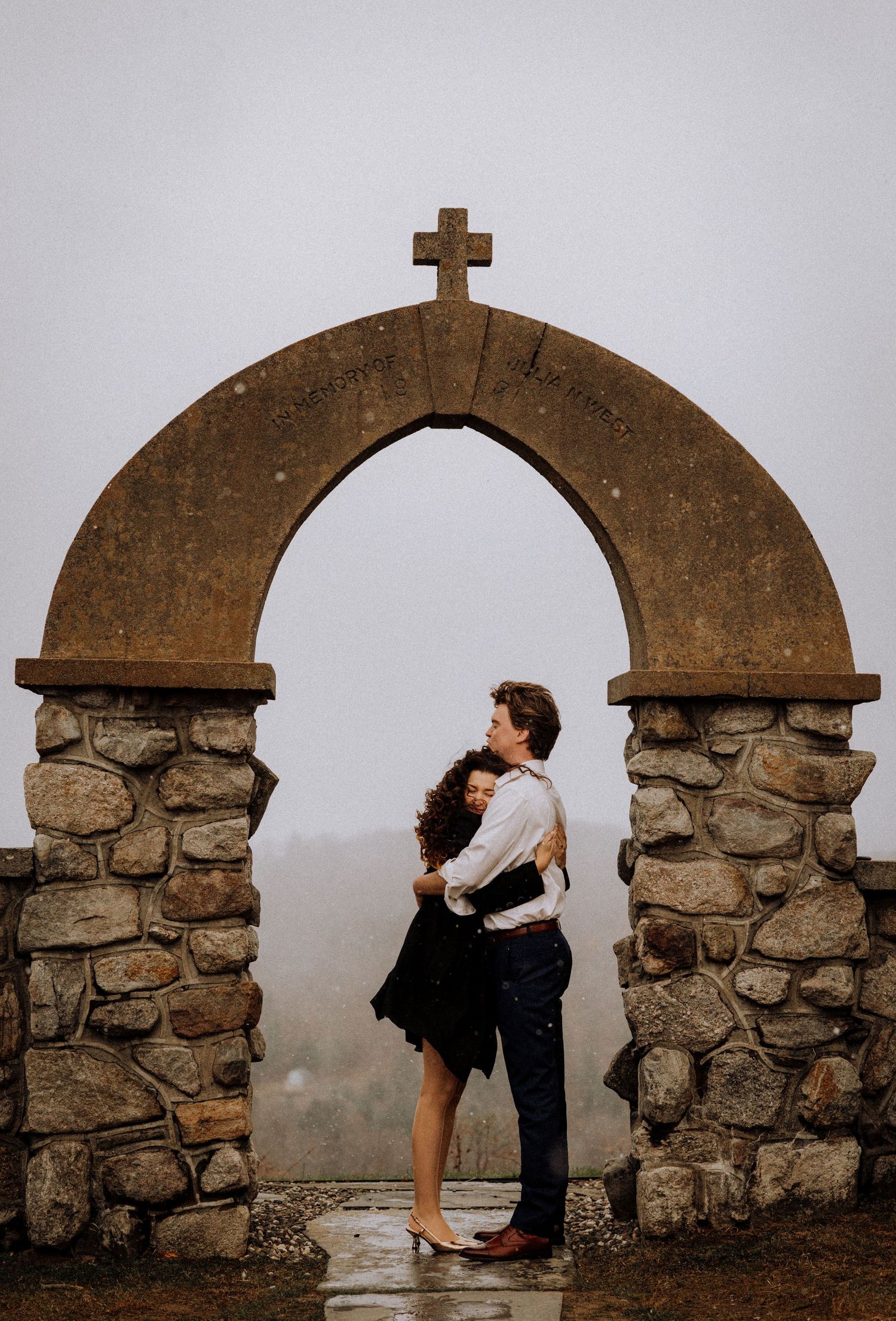 A couple poses between stone archways with a cross on top during a romantic photoshoot.