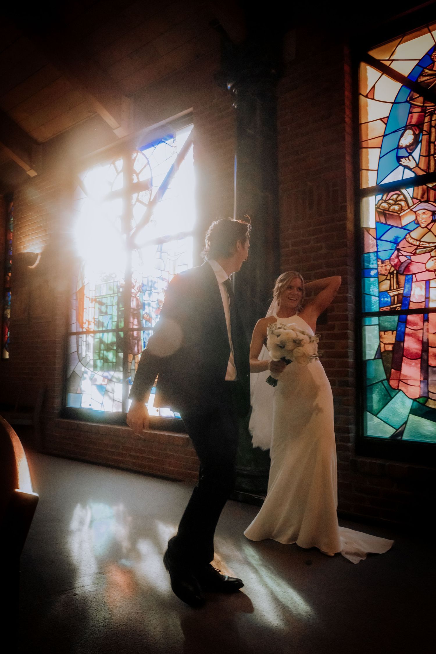 A couple stands in silhouette against dramatic stained glass windows in a church during their wedding ceremony.