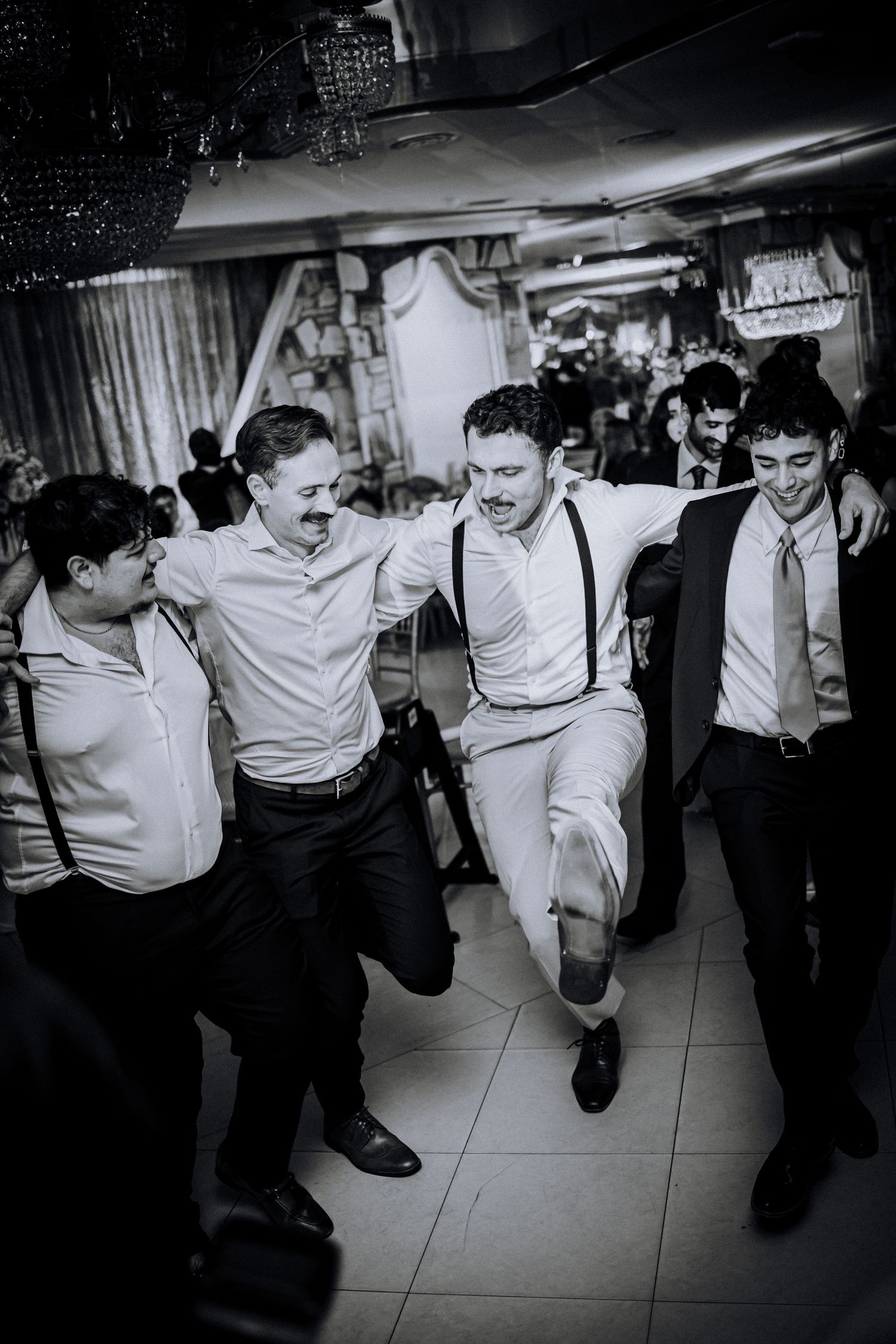A black and white photo of well-dressed men in suits and suspenders dancing and celebrating at a party or reception.