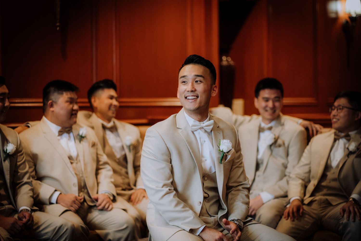 Groomsmen in light beige suits seated in a row during wedding festivities.