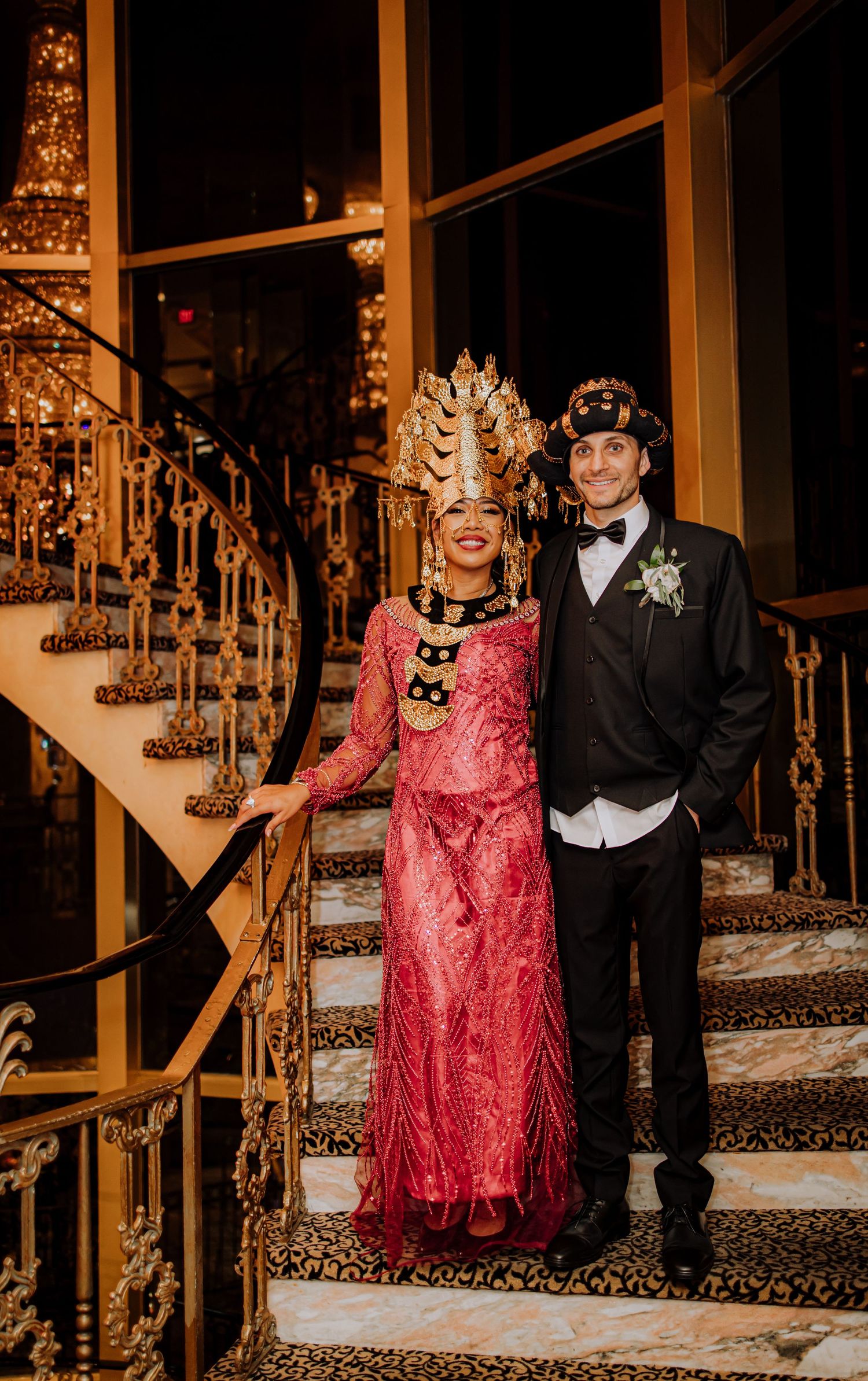 Elegantly dressed figures pose on ornate carpeted stairs with gold railings in formal evening attire.