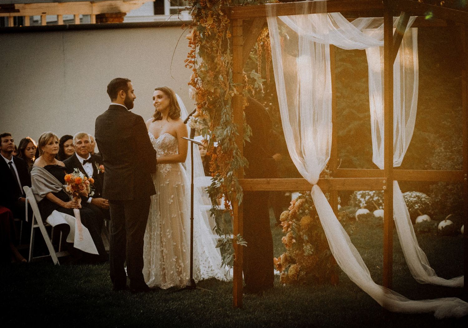 Wedding ceremony setup at sunset with white fabric draping and floral arrangements in a garden setting.