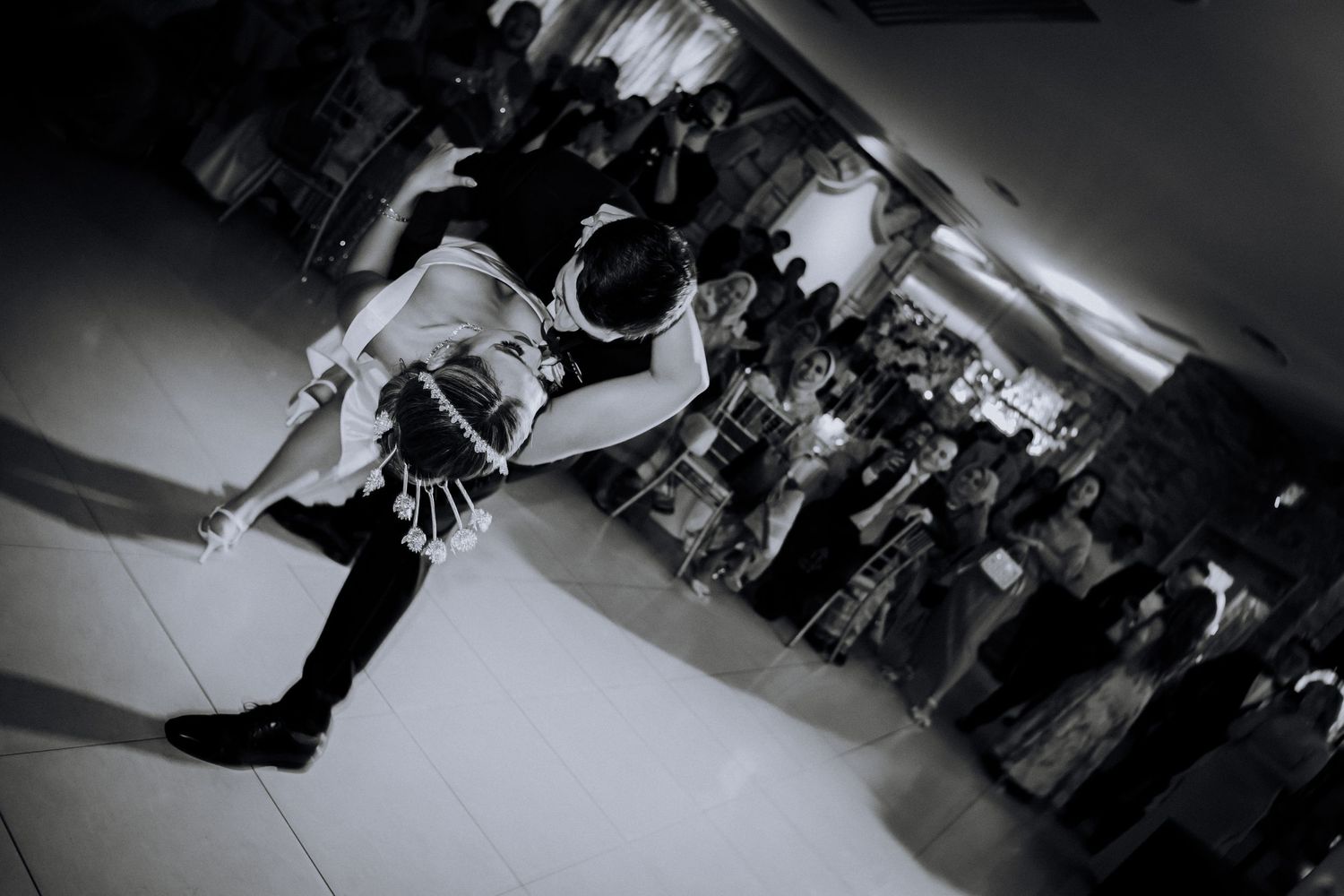 A black and white photo of a couple sharing their first dance at a wedding reception as guests look on.
